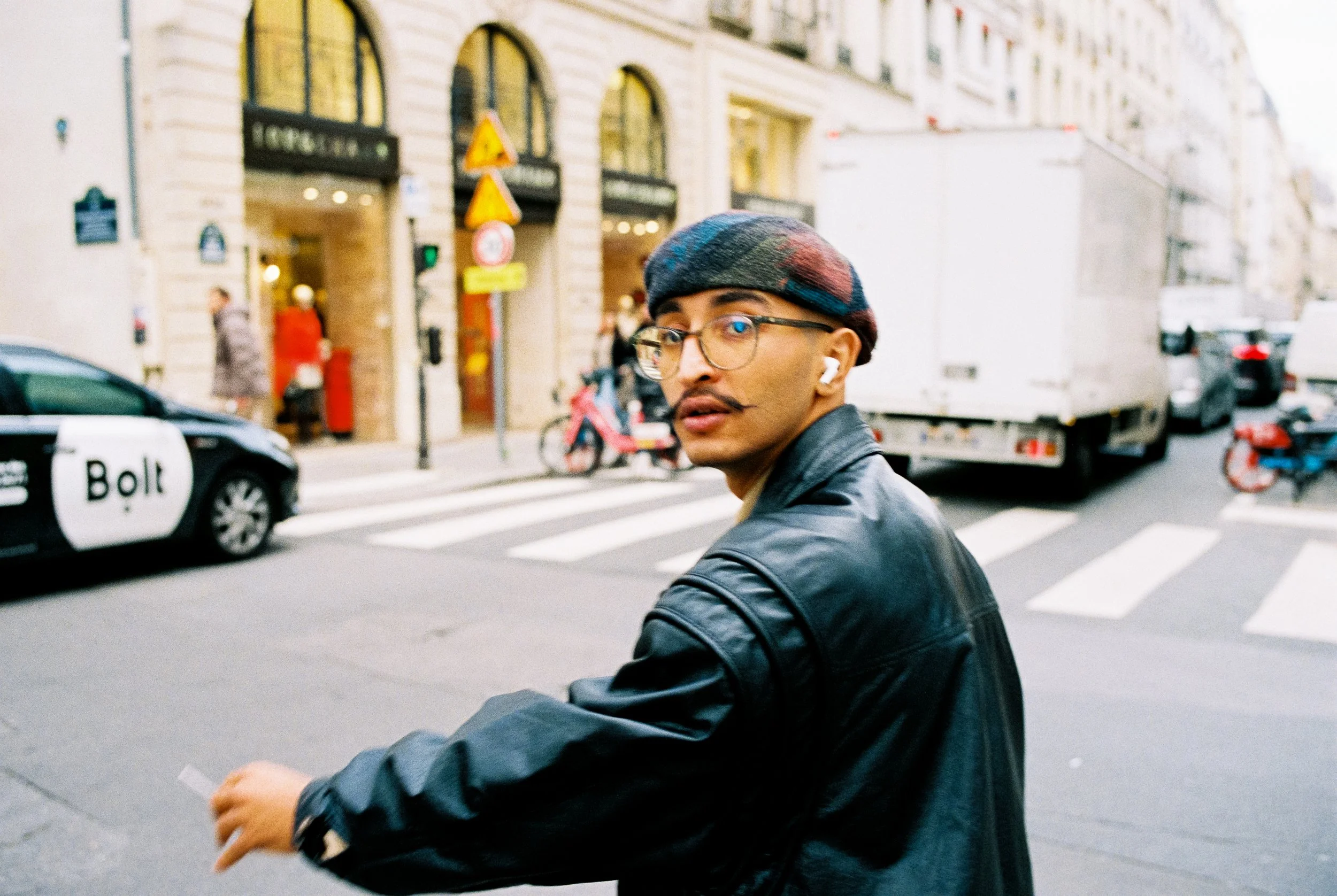 Junger Mann mit Gläsern, Kinnbart und Hörern auf der Straße, im Hintergrund Autos, Menschen und Gebäude, Überquerungshilfe sichtbar.