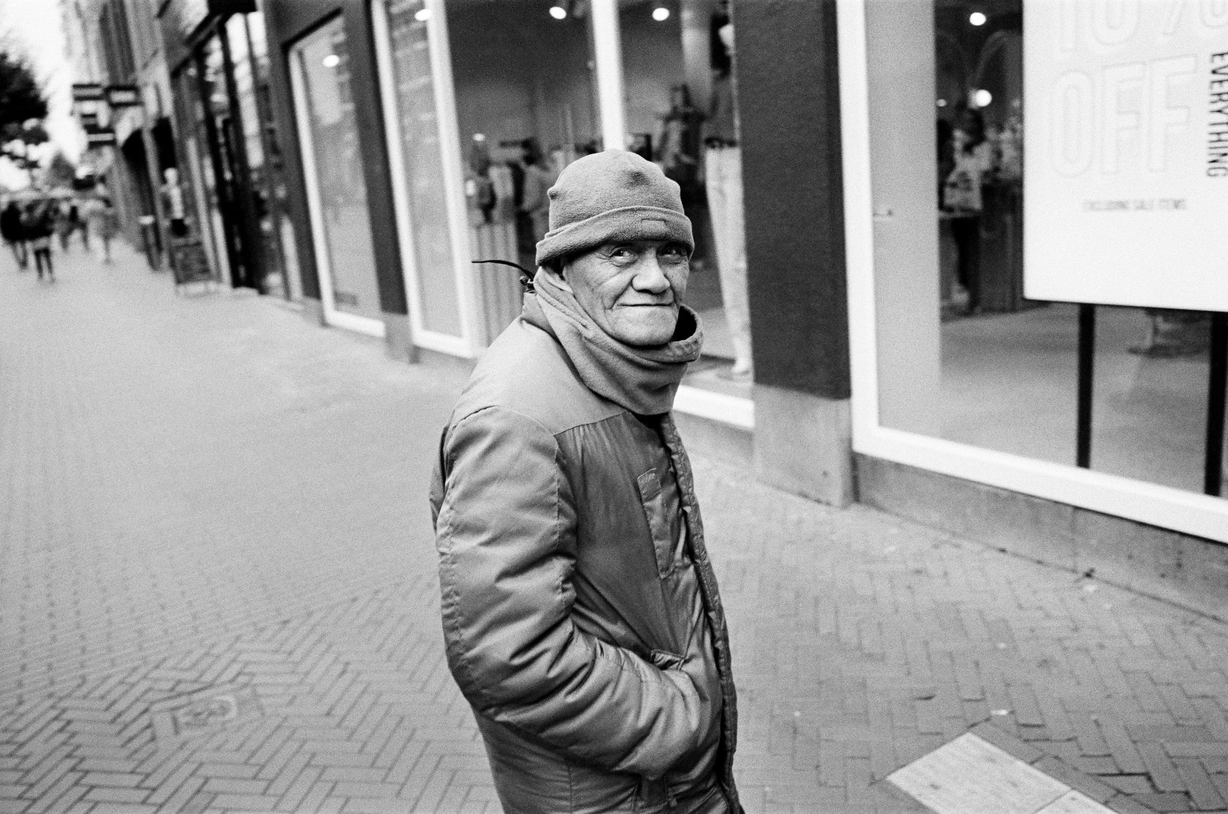 Ein älterer Mann in einem Mantel, Mütze und Schal steht auf einem Bürgersteig in einer Stadt, blickt in die Kamera und hat die Hände in den Taschen.