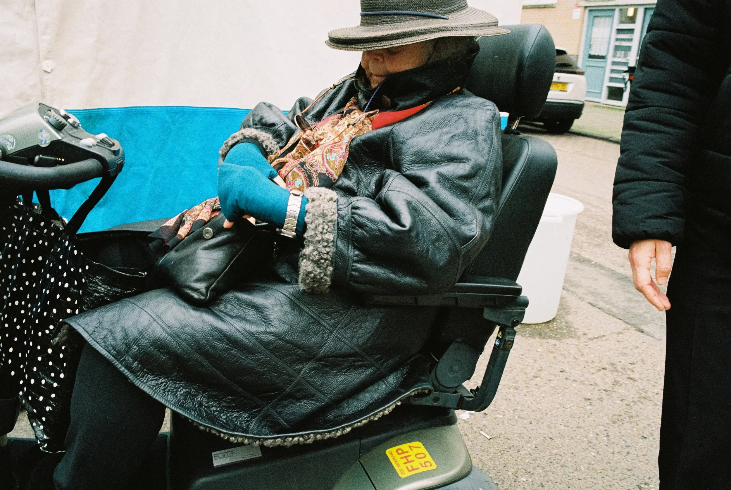 Eine ältere Frau sitzt in einem Elektromobil, trägt einen Hut, eine Lederjacke und hat eine gefütterte Handtasche auf dem Schoß.