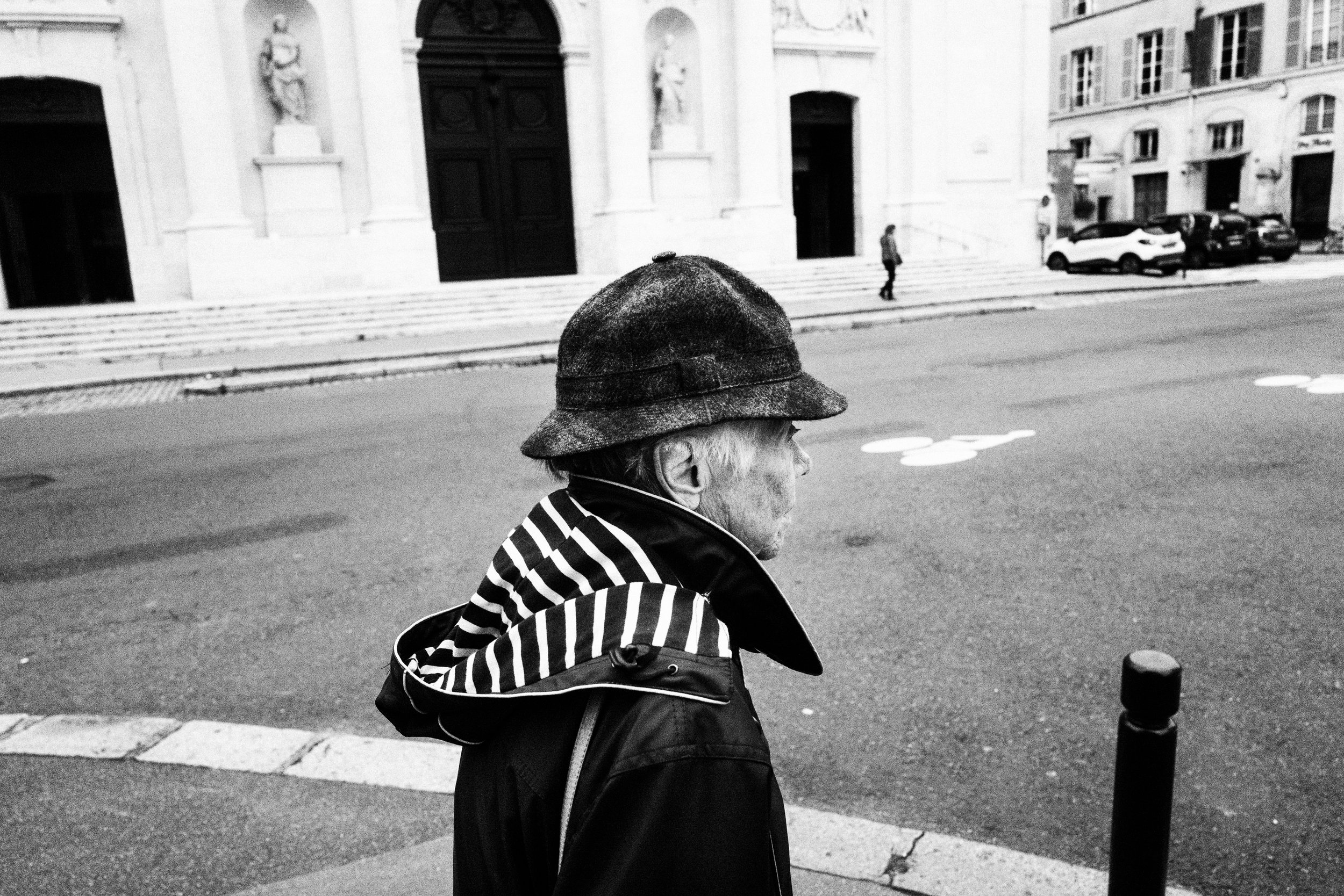 Ein älterer Mann mit grauem Haar trägt einen Hut und eine Jacke mit Streifenmuster. Er steht am Straßenrand vor einem historischen Gebäude in einer Stadt. Das Bild ist schwarz-weiß.