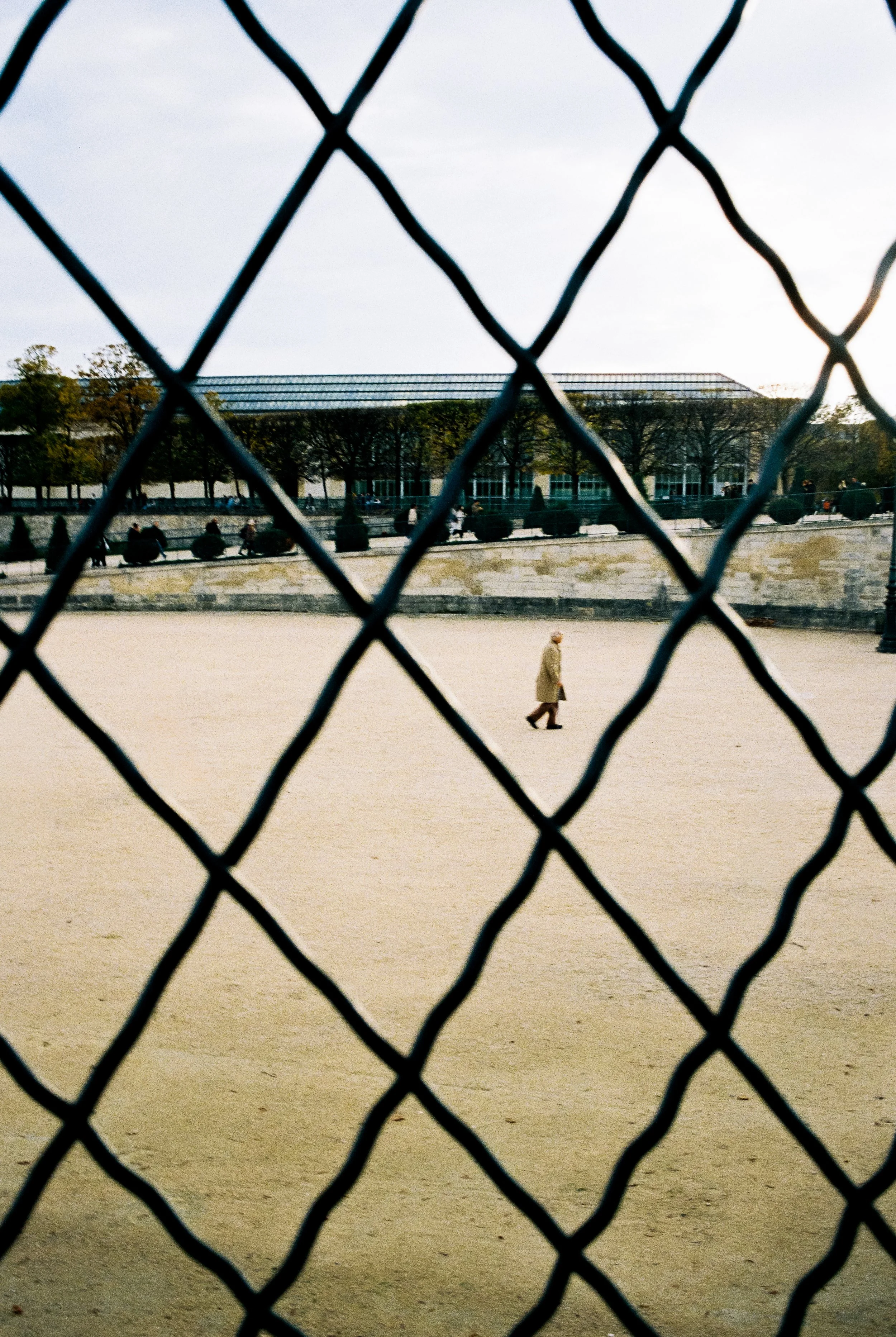 Ein älterer Mensch geht in einem offenen, sandigen Bereich, der durch ein Metallgitter vom Betrachter getrennt ist, vermutlich ein Tennis- oder Basketballplatz.