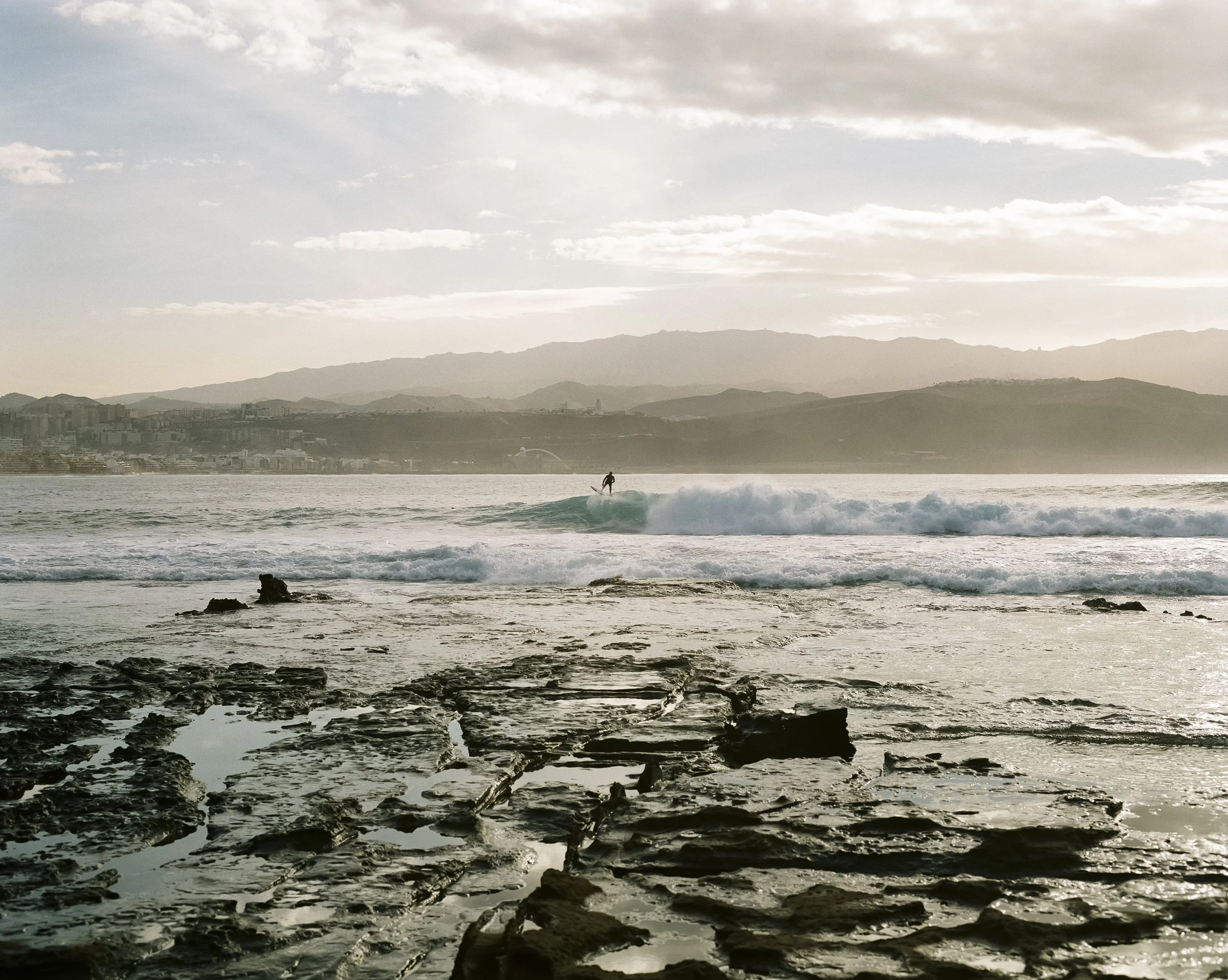 Sonnenuntergang am Meer mit einem Surfer auf einer Welle, im Hintergrund Berge und eine Stadt