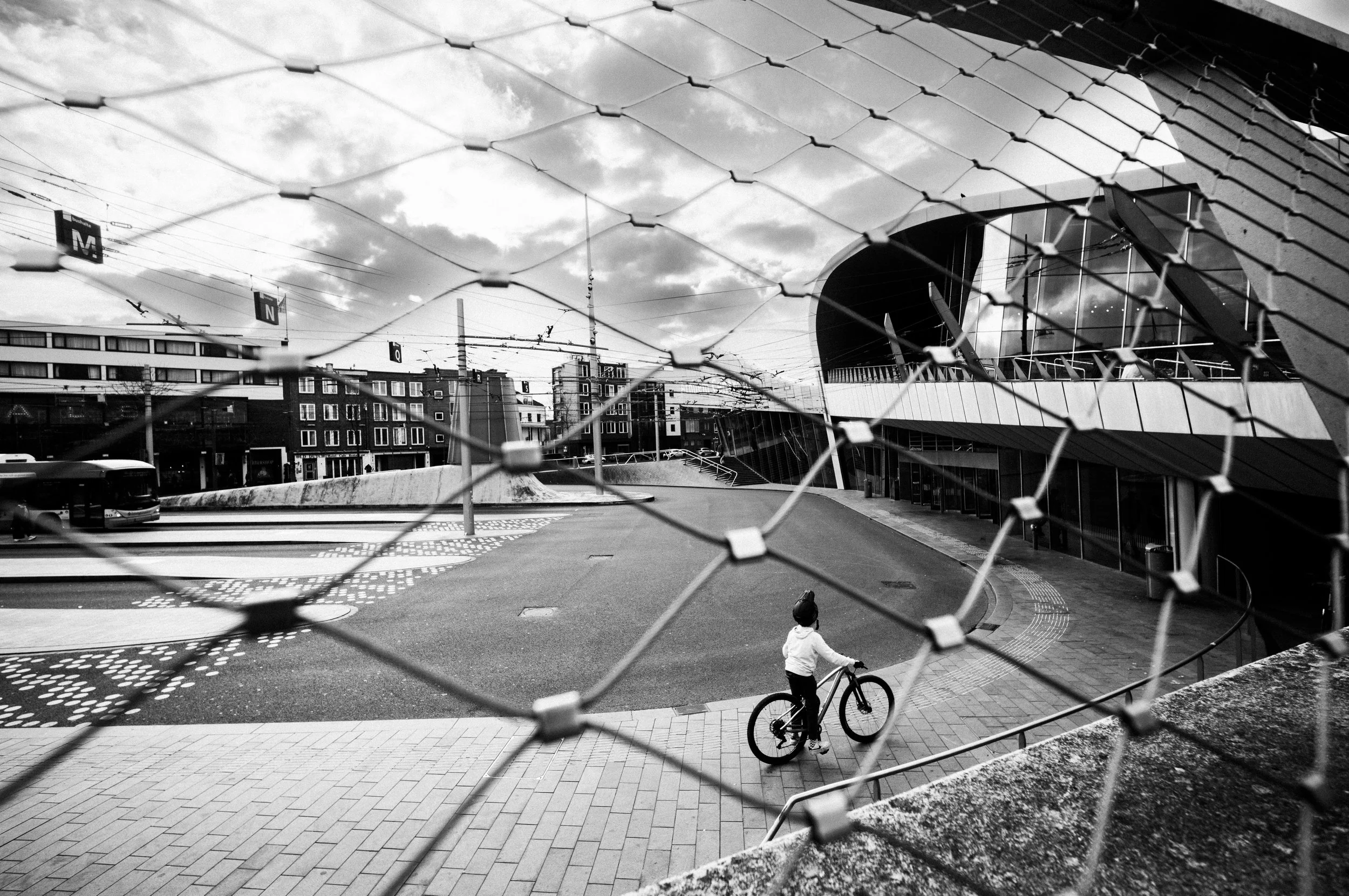 Ein Kind mit Fahrrad, gesehen durch einen Maschendrahtzaun, in einer urbanen Szene mit modernen Gebäuden und Bus, schwarz-weiß.