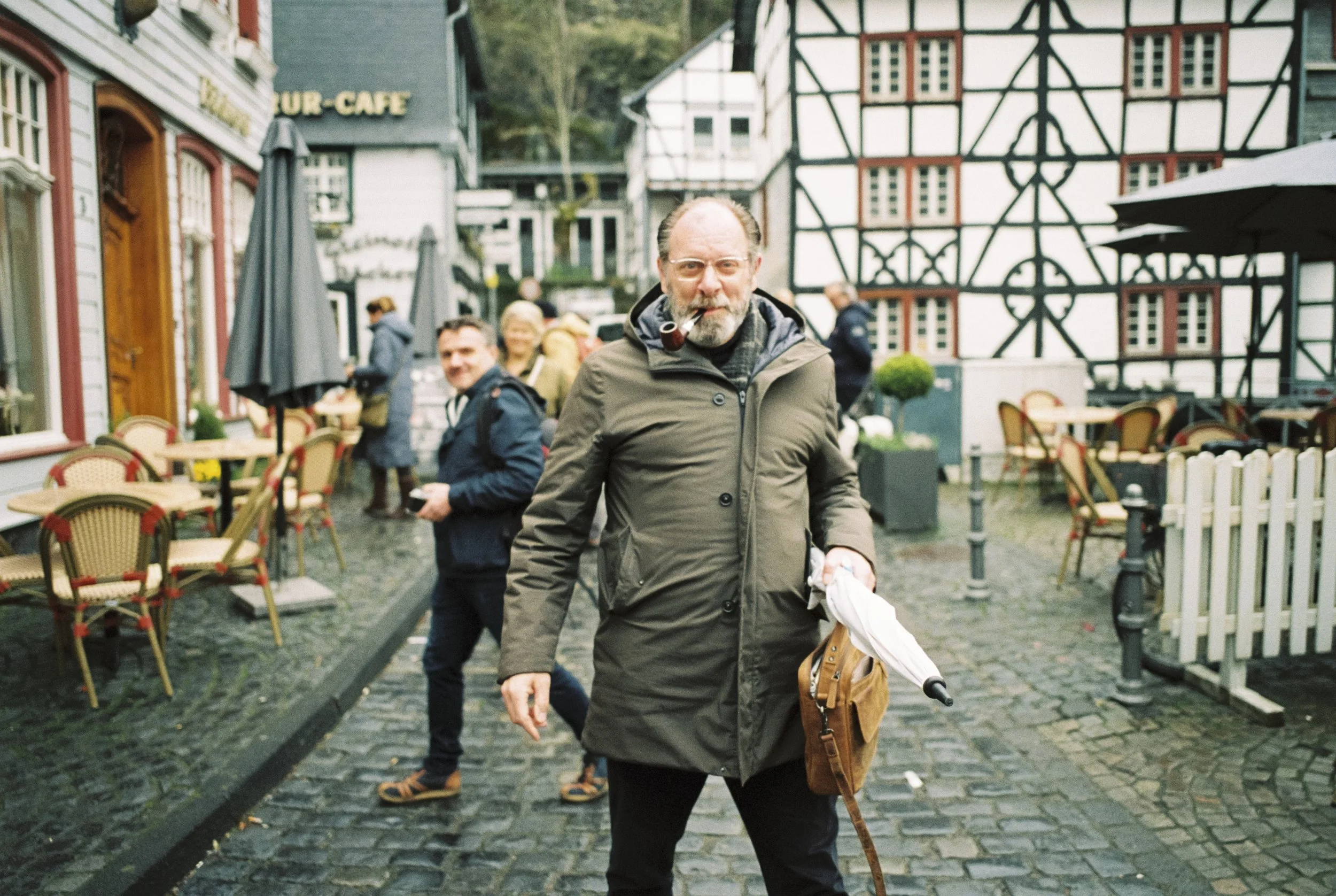 Ein Mann mit Bart und Brille, trägt einen Mantel, raucht eine Pfeife, hält einen Regenschirm in der Hand und steht auf einem gepflasterten Platz vor einem Fachwerkhaus, umgeben von mehreren Personen und Cafés.