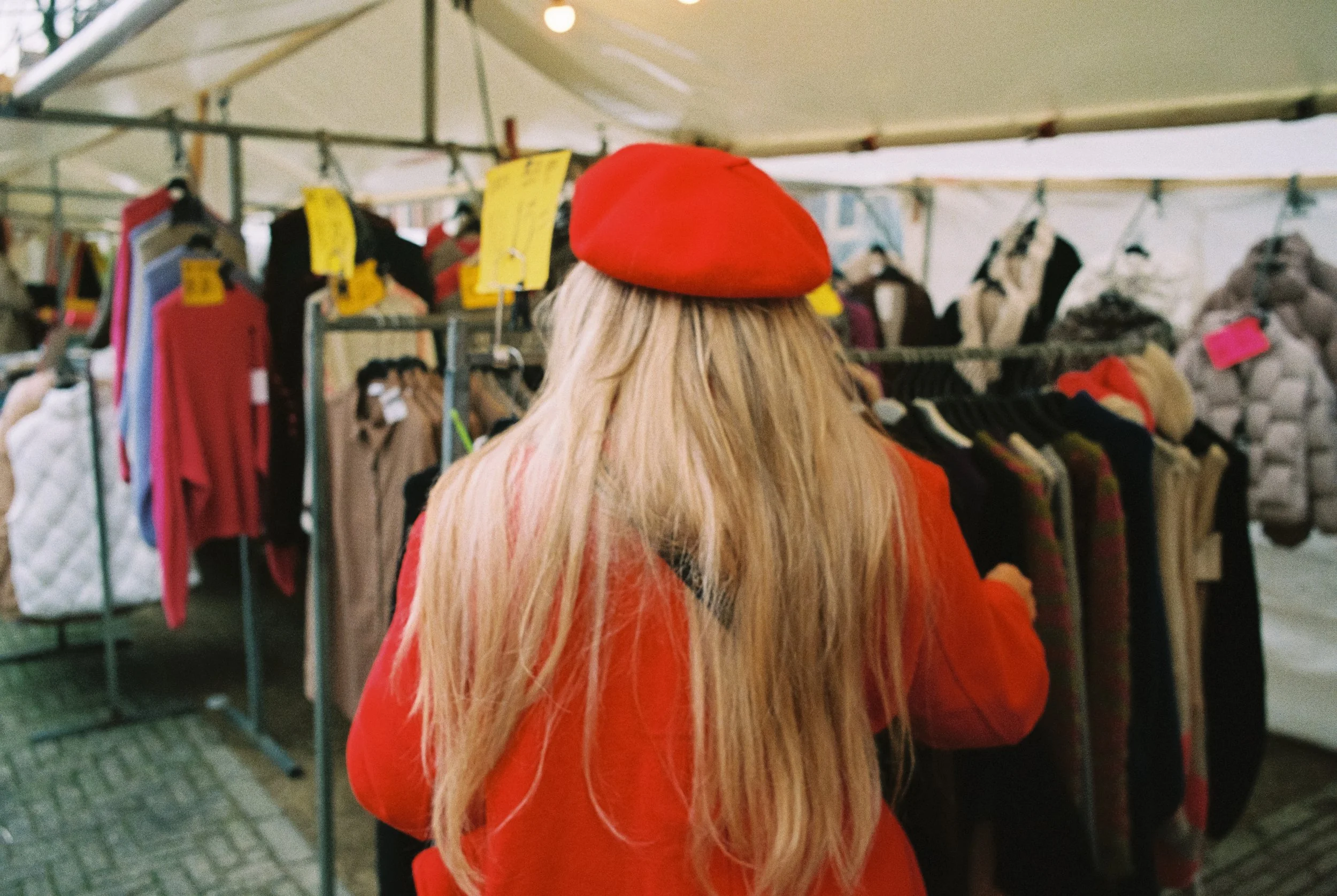 Frau mit blonden Haaren trägt einen roten Hut und einen roten Mantel, steht vor Kleiderständern mit Kleidung auf einem Markt oder Secondhand-Laden.