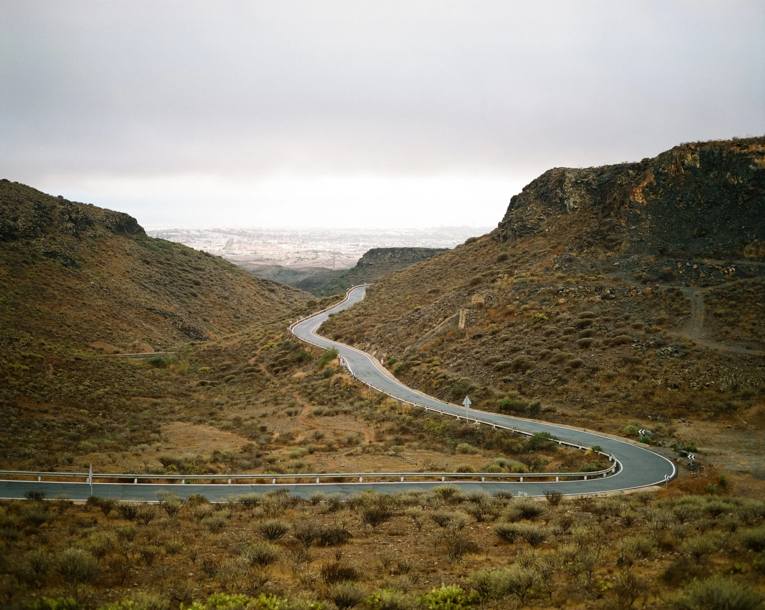 Eine kurvige Straße in einer bergigen, trockenen Landschaft unter einem bewölkten Himmel.