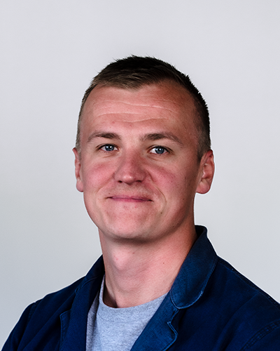 A young man with short brown hair, blue eyes, and a slight smile, wearing a navy blue shirt and a gray t-shirt underneath, posing against a plain gray background.