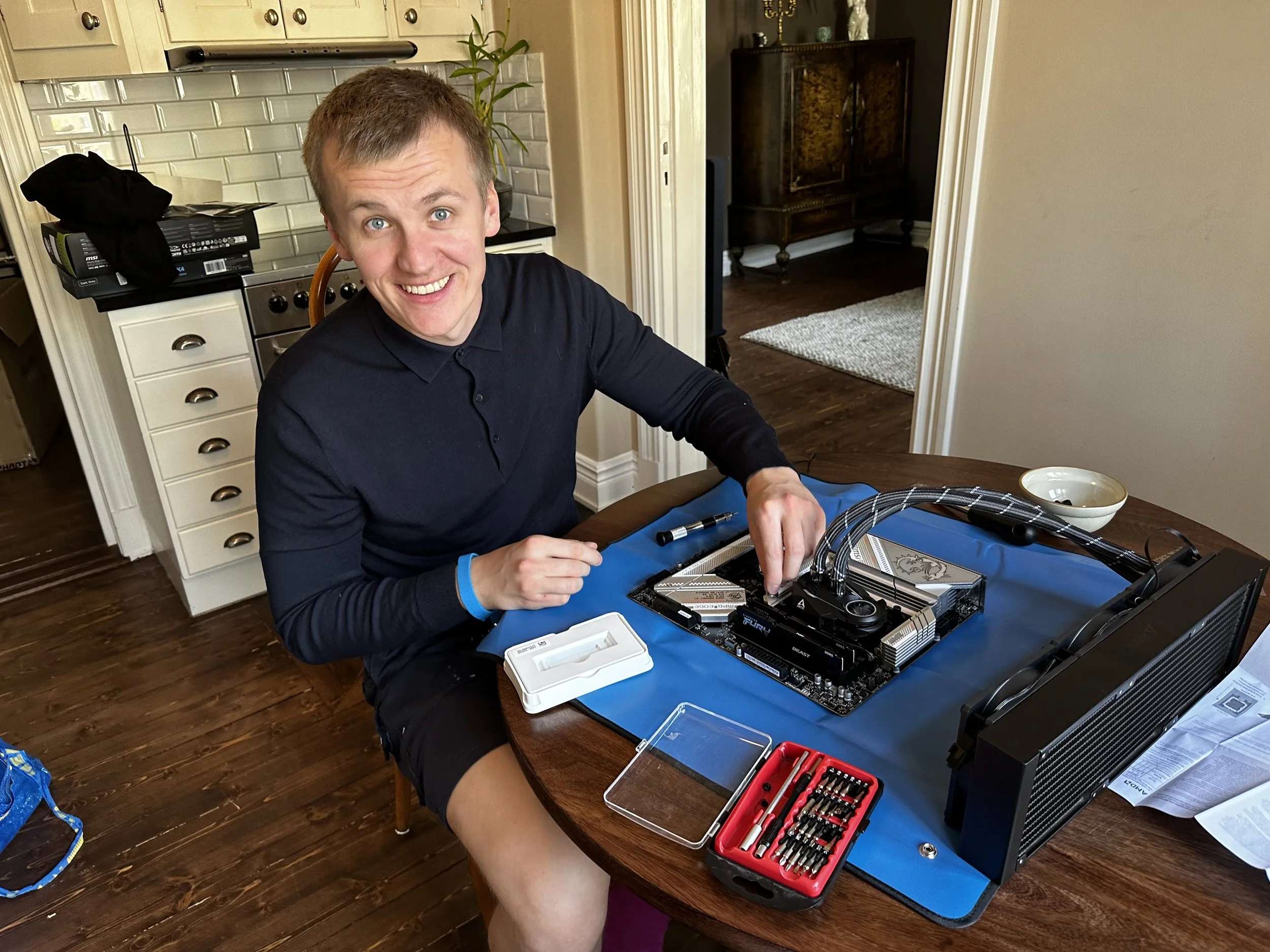 A young man with short brown hair and blue eyes, wearing a black long-sleeve shirt and black shorts, is sitting at a round wooden table assembling a computer. He's smiling and looking at the camera. On the table, there are various computer components, tools, and a blue mat, with a kitchen and hallway visible in the background.