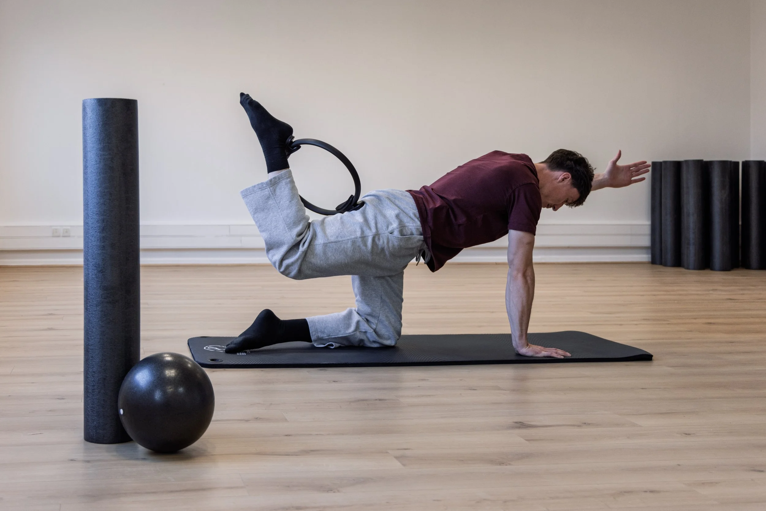 Hommes faisant du yoga sur un tapis dans une pièce vide, avec un rouleau de foam et un ballon en face.