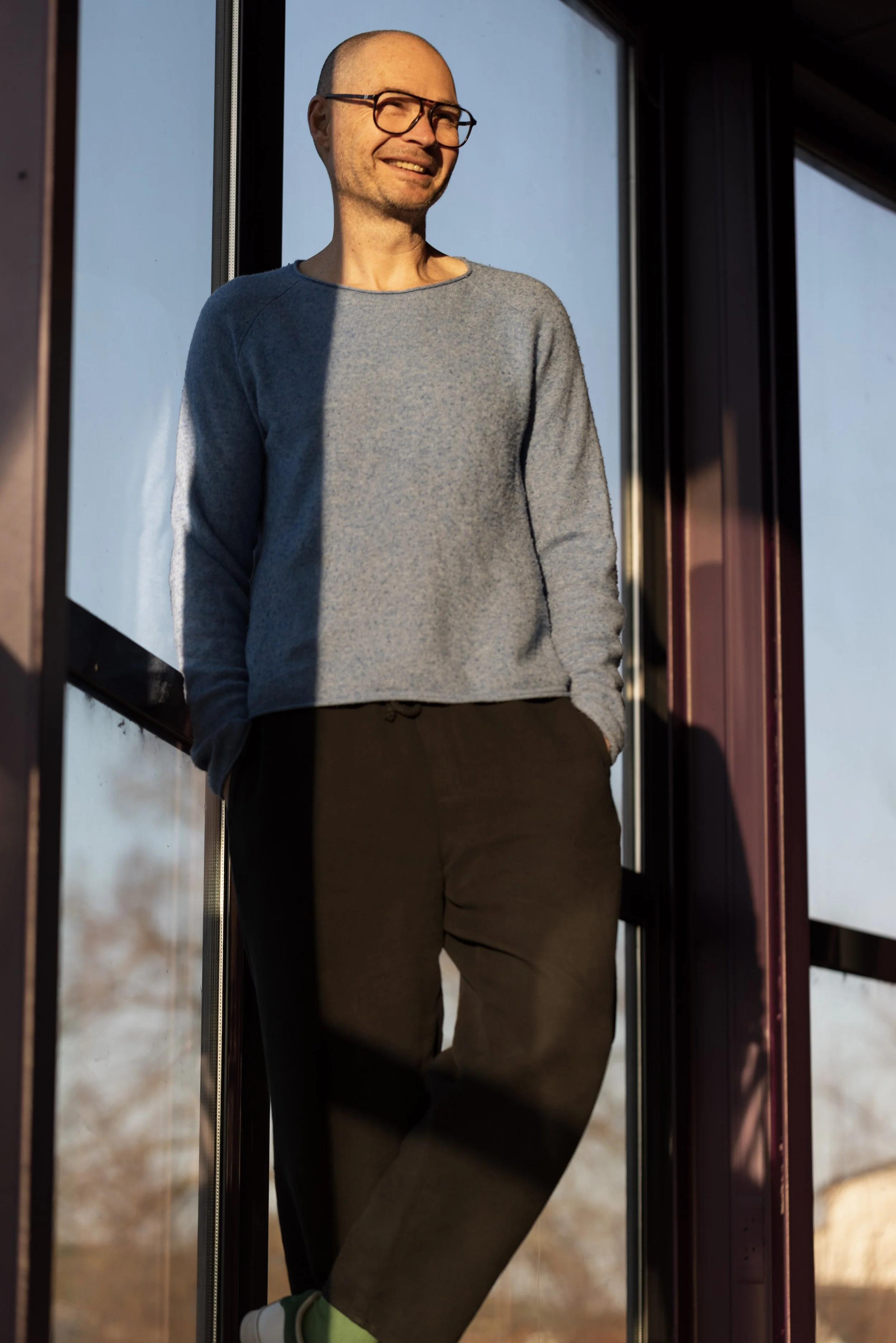 Un homme souriant, portant des lunettes, vêtue d'un sweat gris et de pantalons noirs, se tient dans une structure en verre, avec un ciel bleu en arrière-plan.