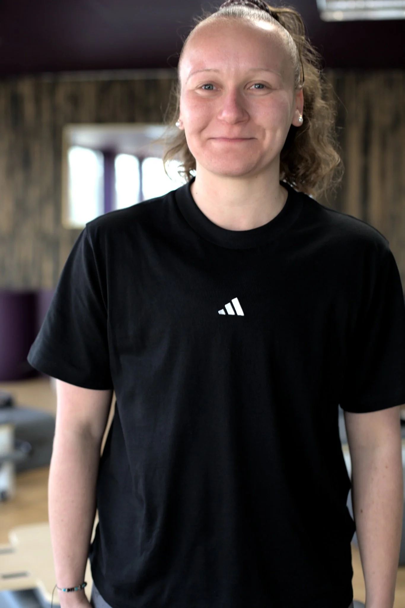 Une femme souriante portant un t-shirt noir avec un logo blanc, dans un intérieur avec des murs en bois et des fenêtres à l'arrière-plan.