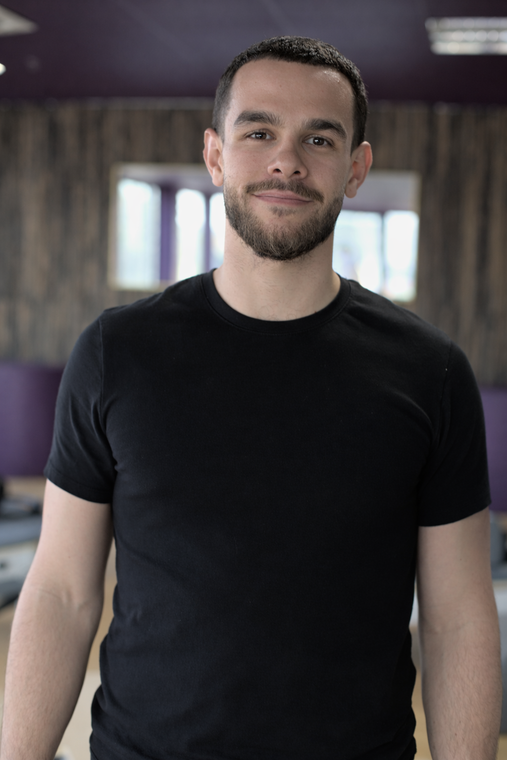 Un jeune homme avec une barbe, portant un t-shirt noir, souriant doucement, dans un intérieur avec fenêtres et murs en bois.