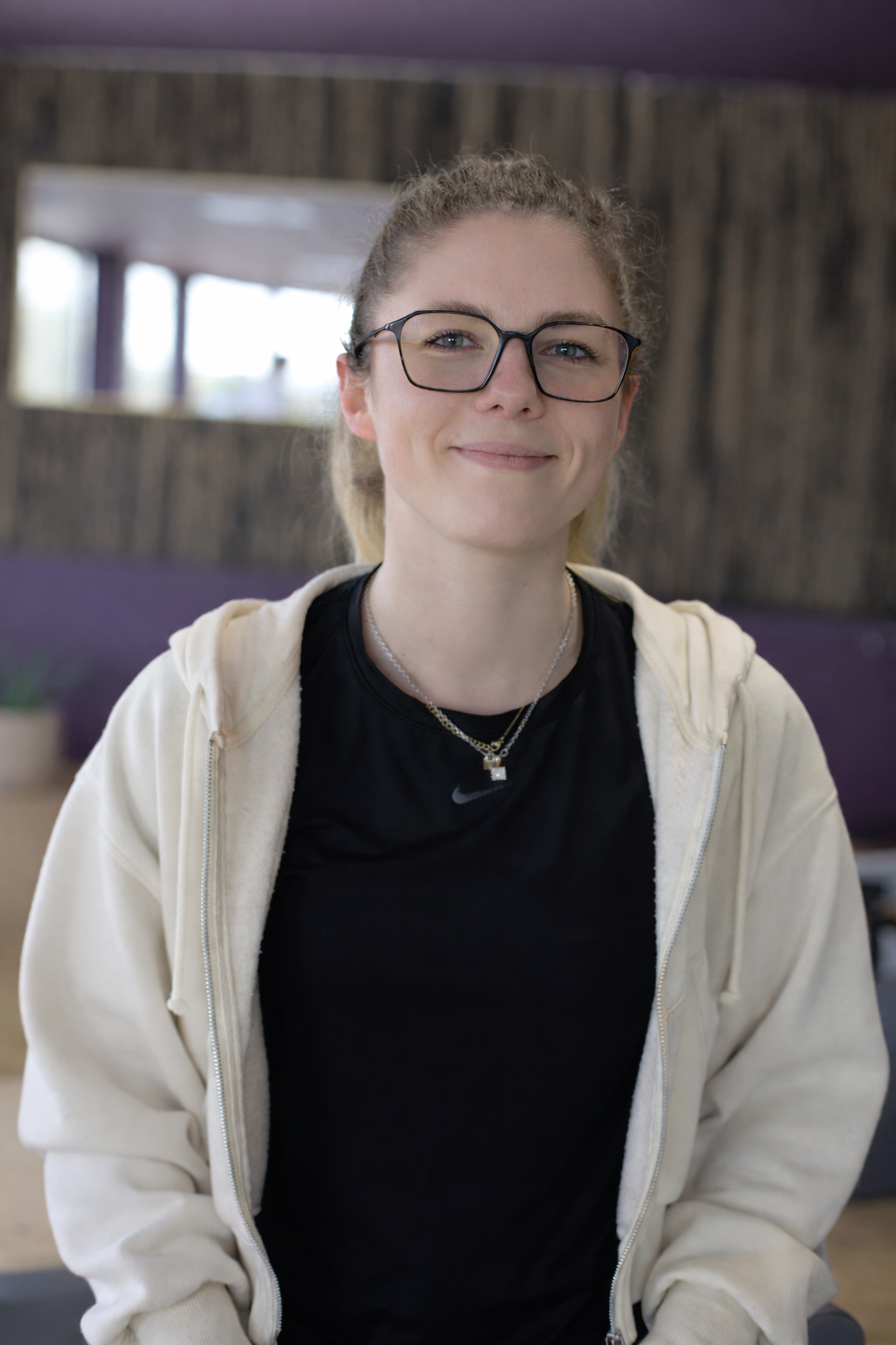 Jeune femme souriante avec des lunettes, portant un t-shirt noir Nike et un sweat crème, dans un intérieur moderne avec décoration en bois et plantes.