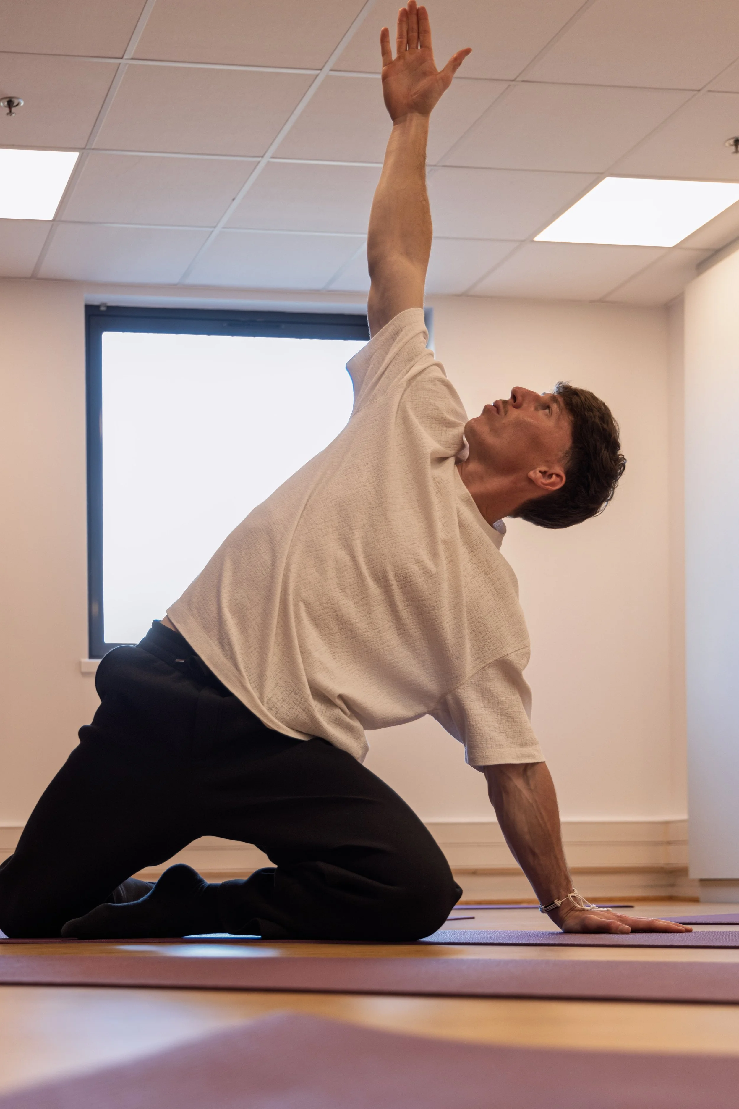 Un homme fait du yoga dans une salle avec une fenêtre, en position de posture de yoga, appuyé sur un genou avec un bras levé vers le haut.