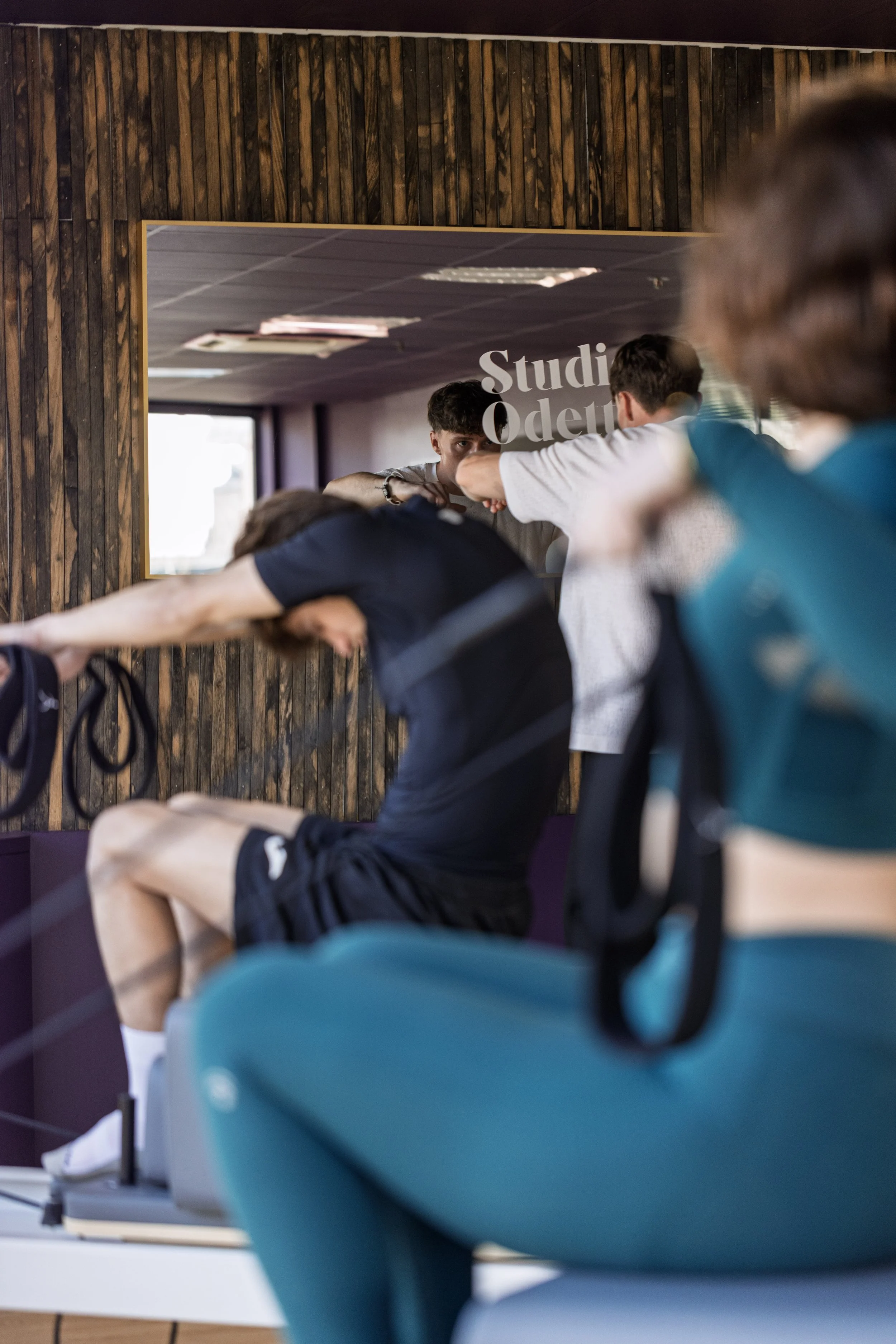 Une séance de Pilates Refomer en groupe dans une salle avec des murs en bois, avec des personnes pliées en avant sur des tapis, regardant vers l'avant.