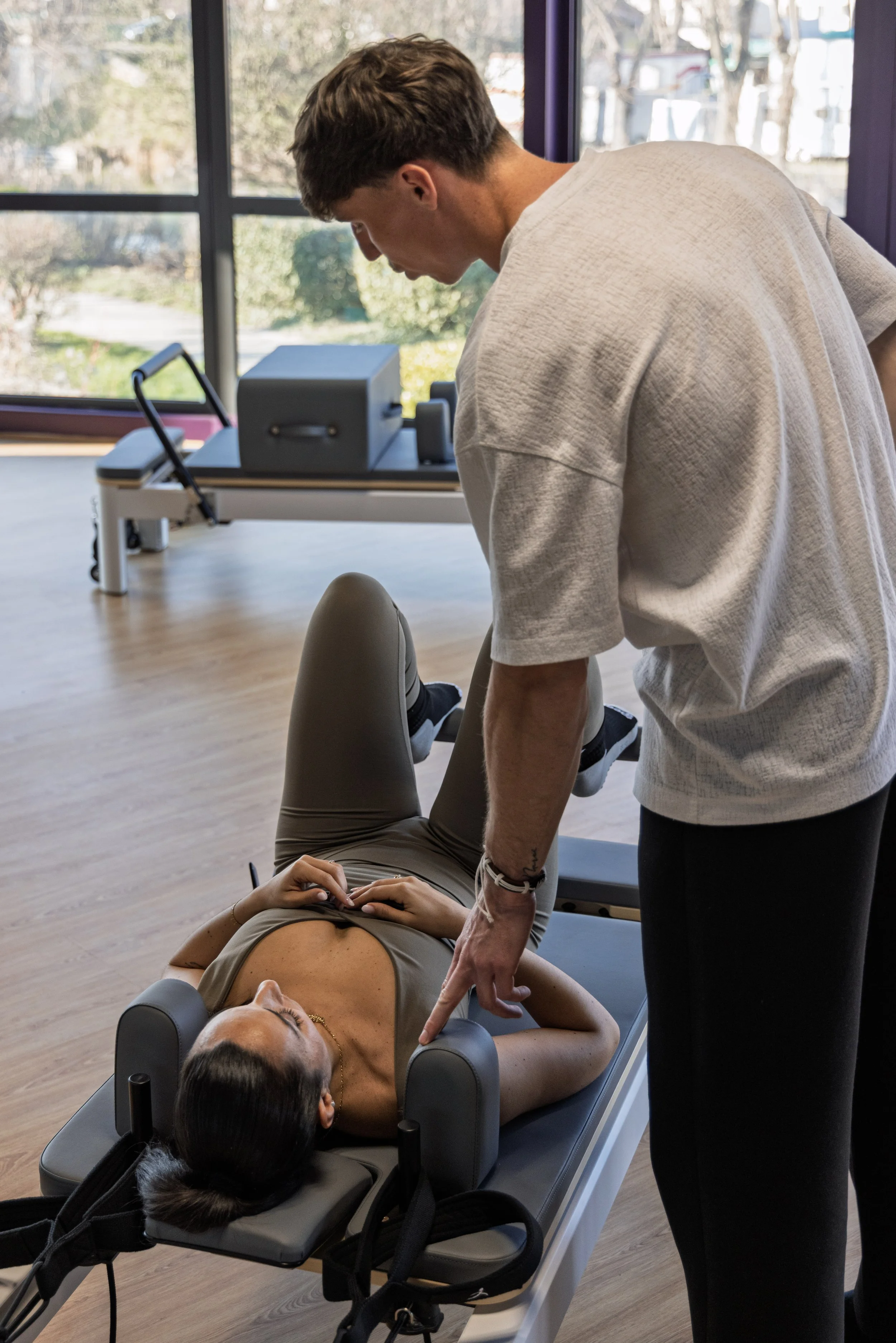 Une femme allongée sur une table de Pilates Reformer, un homme lui donne des conseils sur ça posture, dans une salle avec vue sur le jardin.