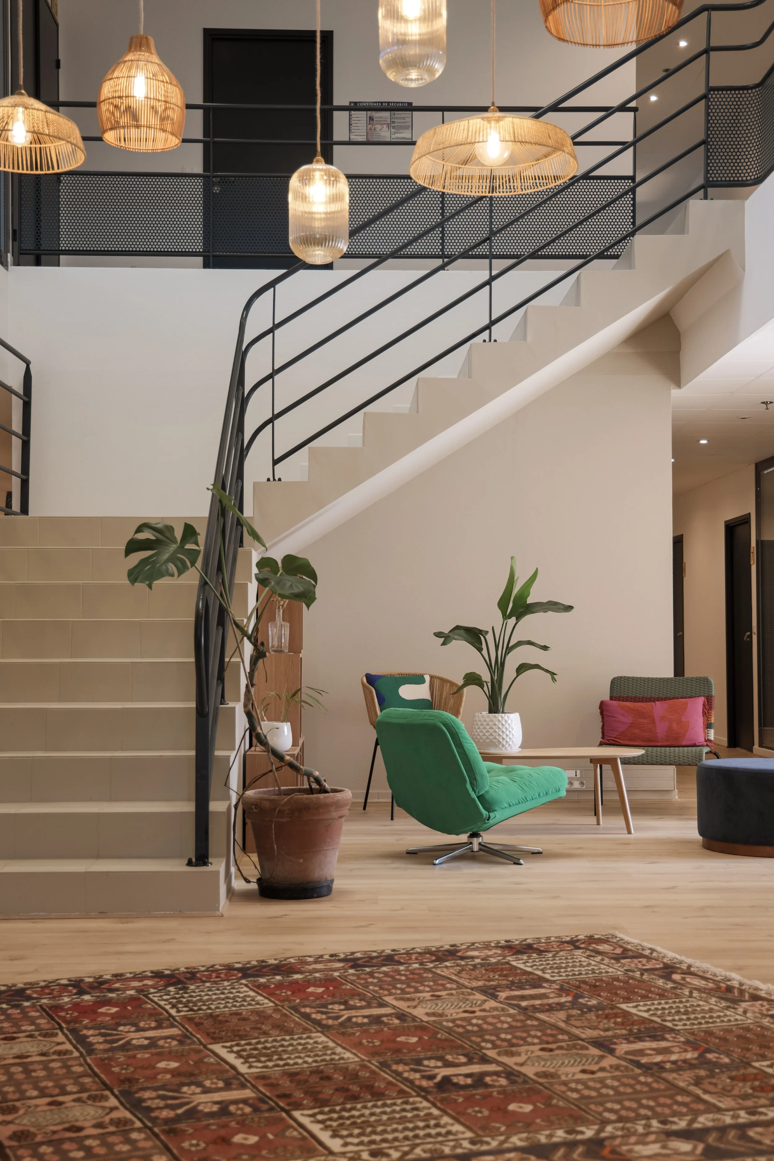 Intérieur d'un hall avec escalier, lampes suspendues en rotin, fauteuils colorés et plantes en pot.