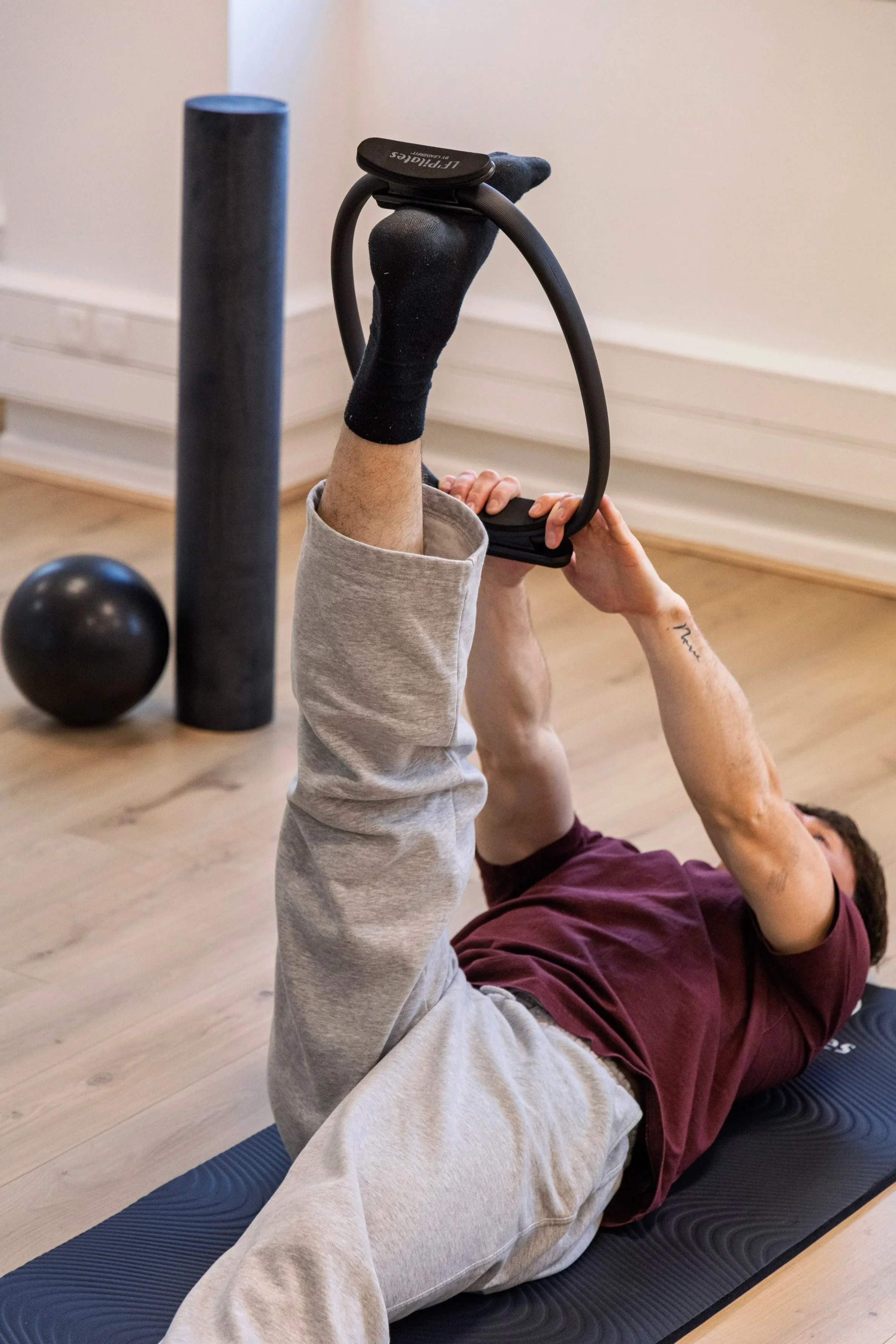 Une personne allongée sur un tapis de yoga, tenant un cerceau de pilates avec ses jambes levées, dans une salle de sport. Deux balles noires et un rouleau de foam noir sont visibles en arrière-plan.