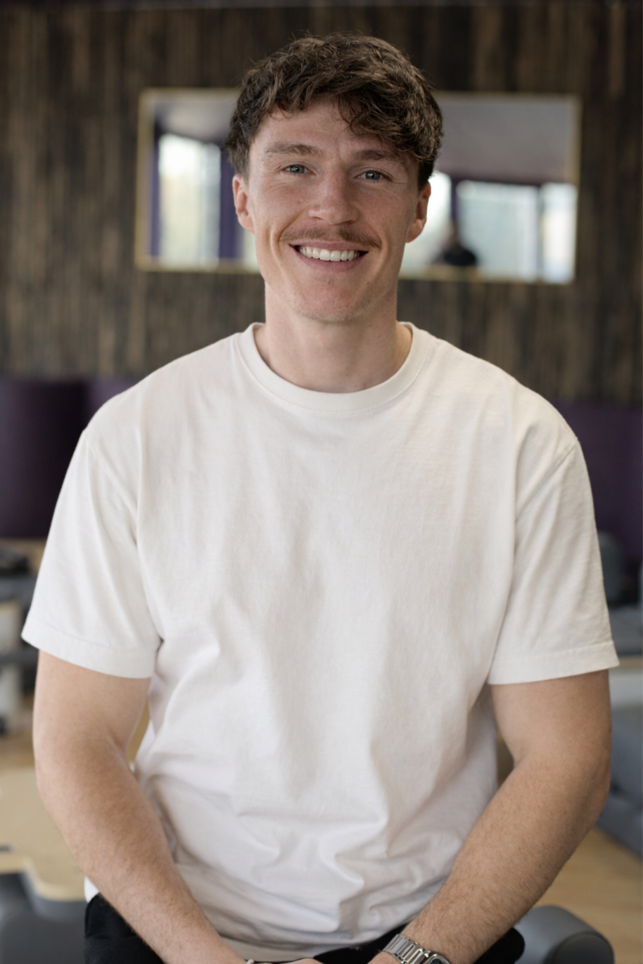 Jeune homme souriant avec t-shirt blanc, assis dans un intérieur moderne en bois, fenêtre en arrière-plan.