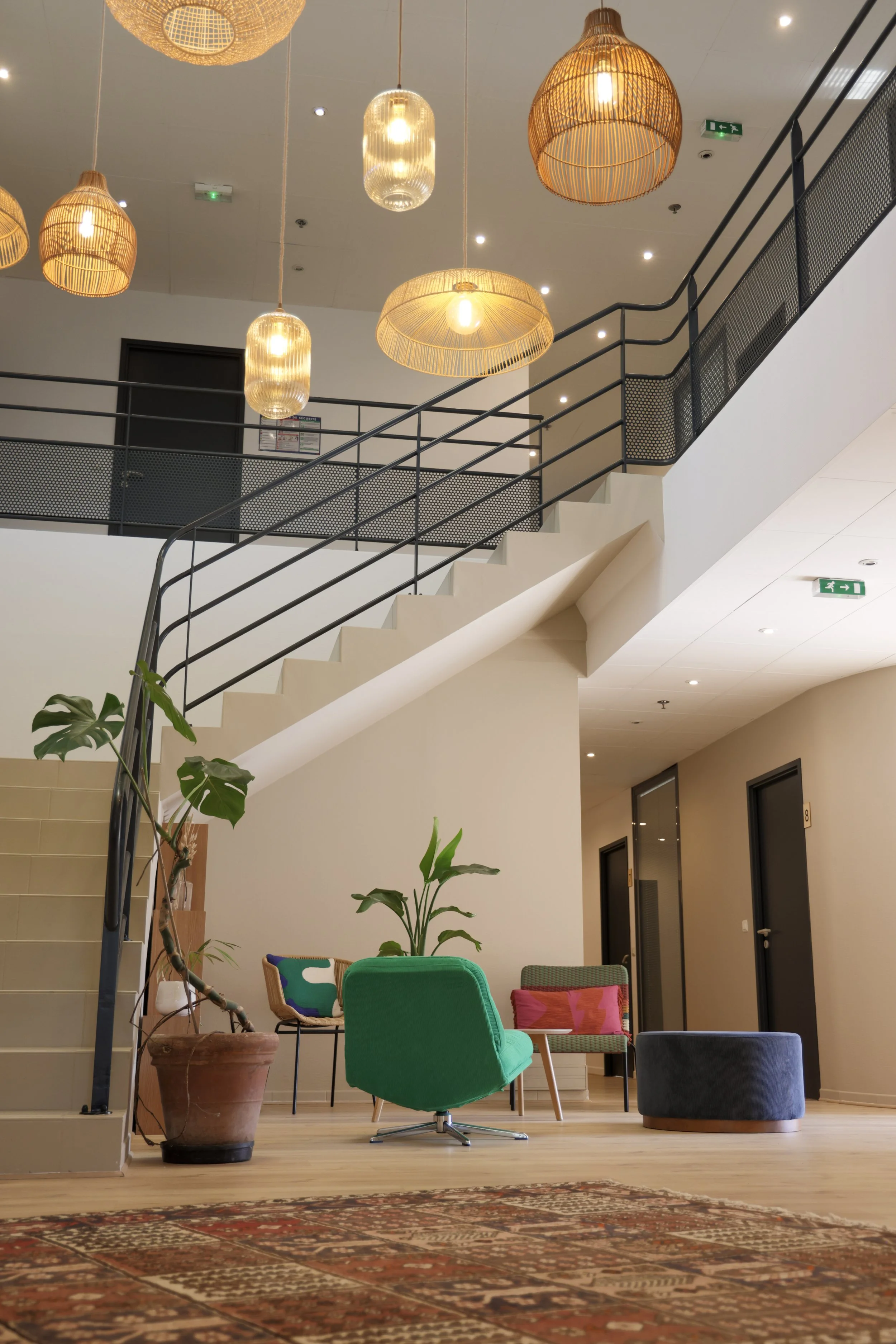 Intérieur moderne d'un bâtiment, avec fauteuils colorés, plantes en pot, escalier en métal, éclairage suspendu en rotin, murs clairs et portes en bois, tapis traditionnel.