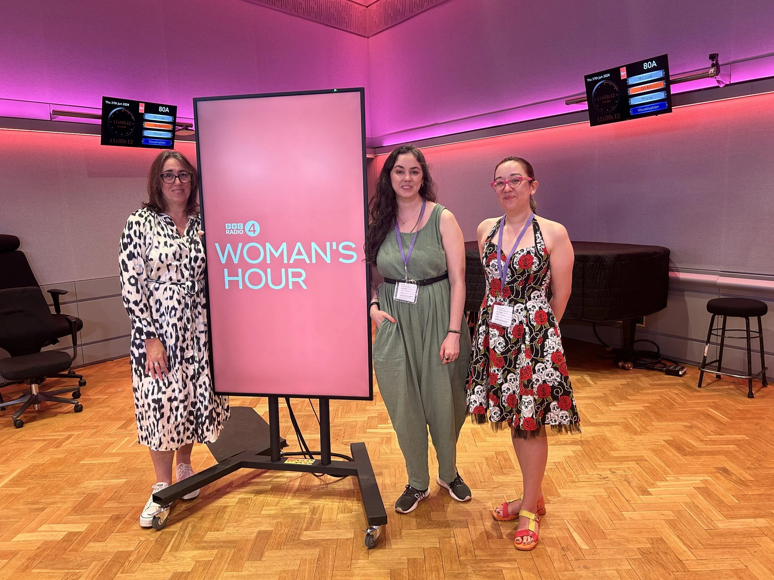Three women standing next to a pink display screen that reads 'BBC Radio 4 Woman's Hour', in a room with a wooden floor and purple lighting. Two screens are mounted on the wall behind them, displaying digital information.