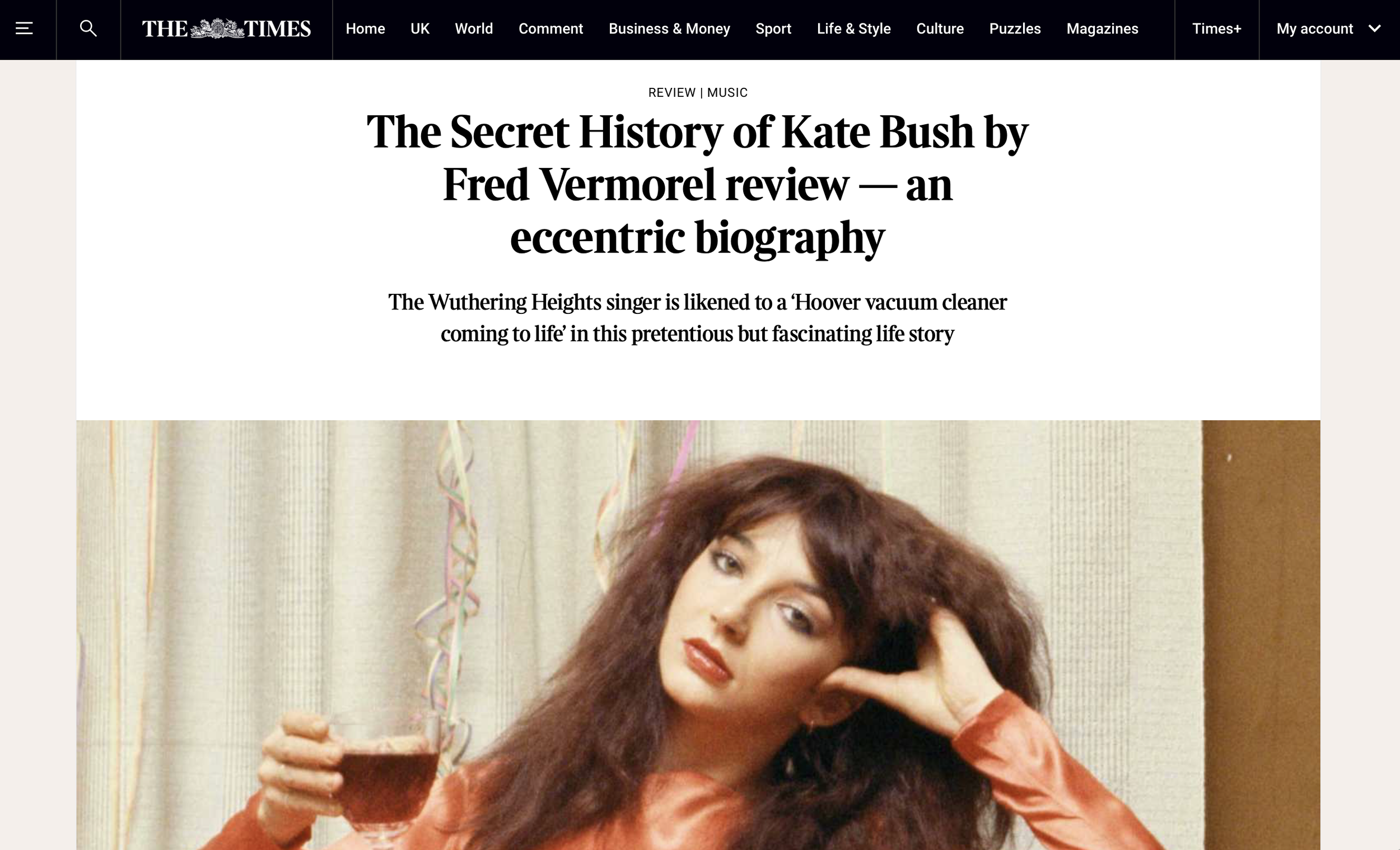 A woman with long curly hair holding a glass of red drink, sitting with a relaxed pose against a beige curtain background with colorful streamers.