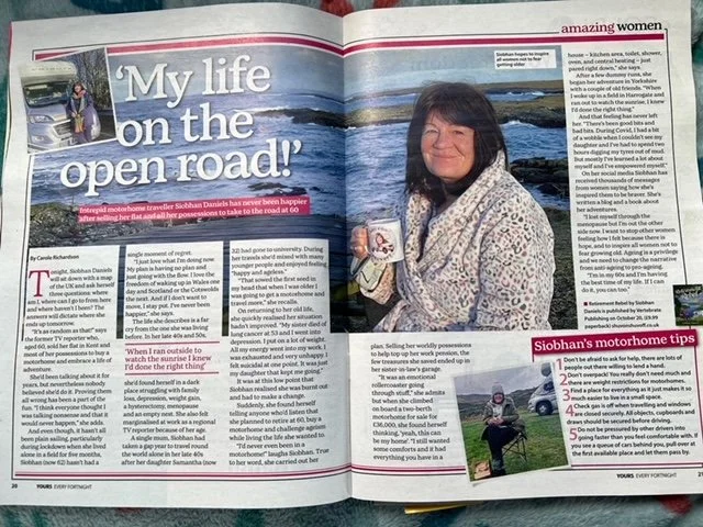 A woman with dark hair and a patterned sweater sitting outside on rocks by the ocean, holding a mug and smiling at the camera.
