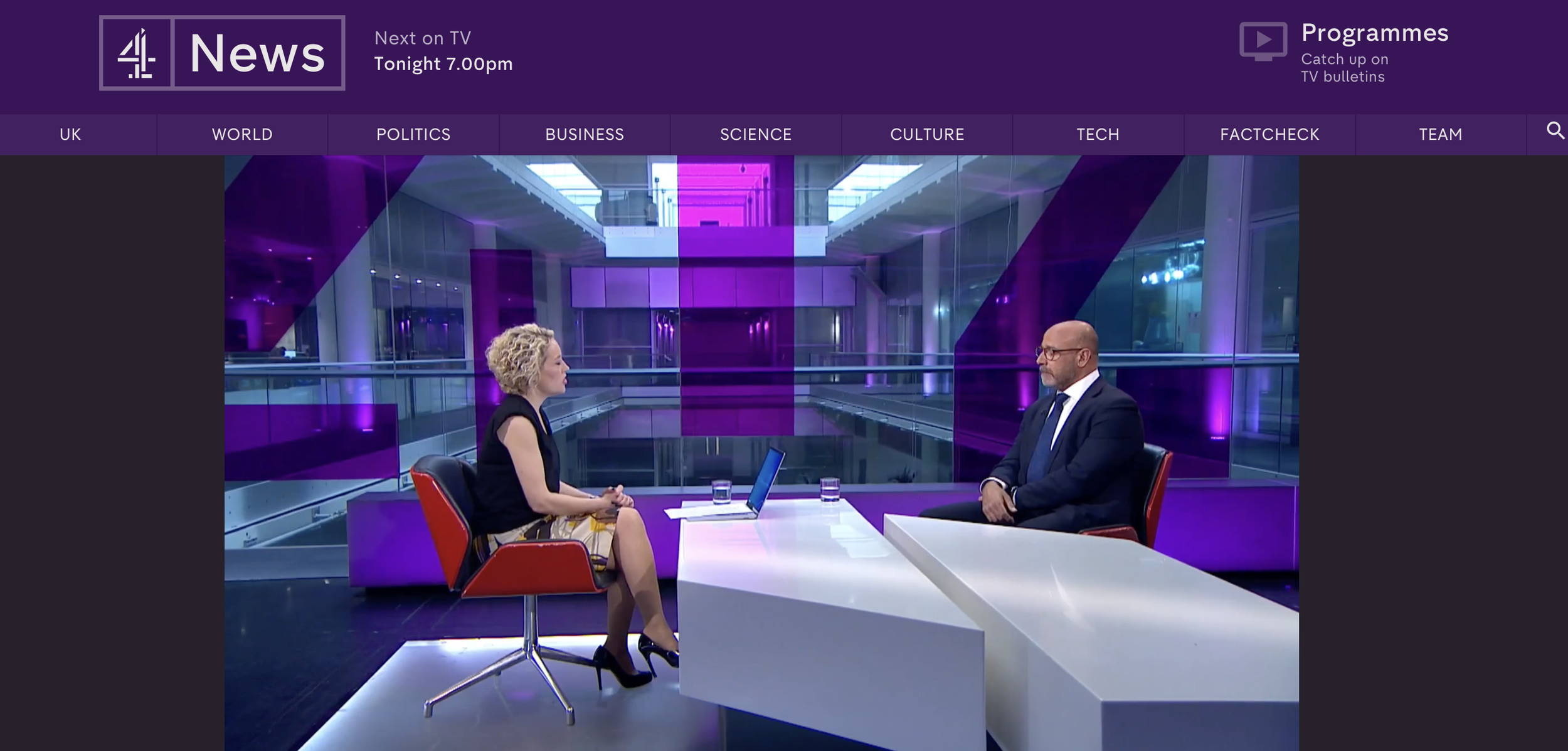 A television news interview taking place in a studio with a modern, purple-lit background. A woman with curly blonde hair and a black top sits on the left, while a man with glasses, a beard, and a dark suit sits on the right. There are two glasses of