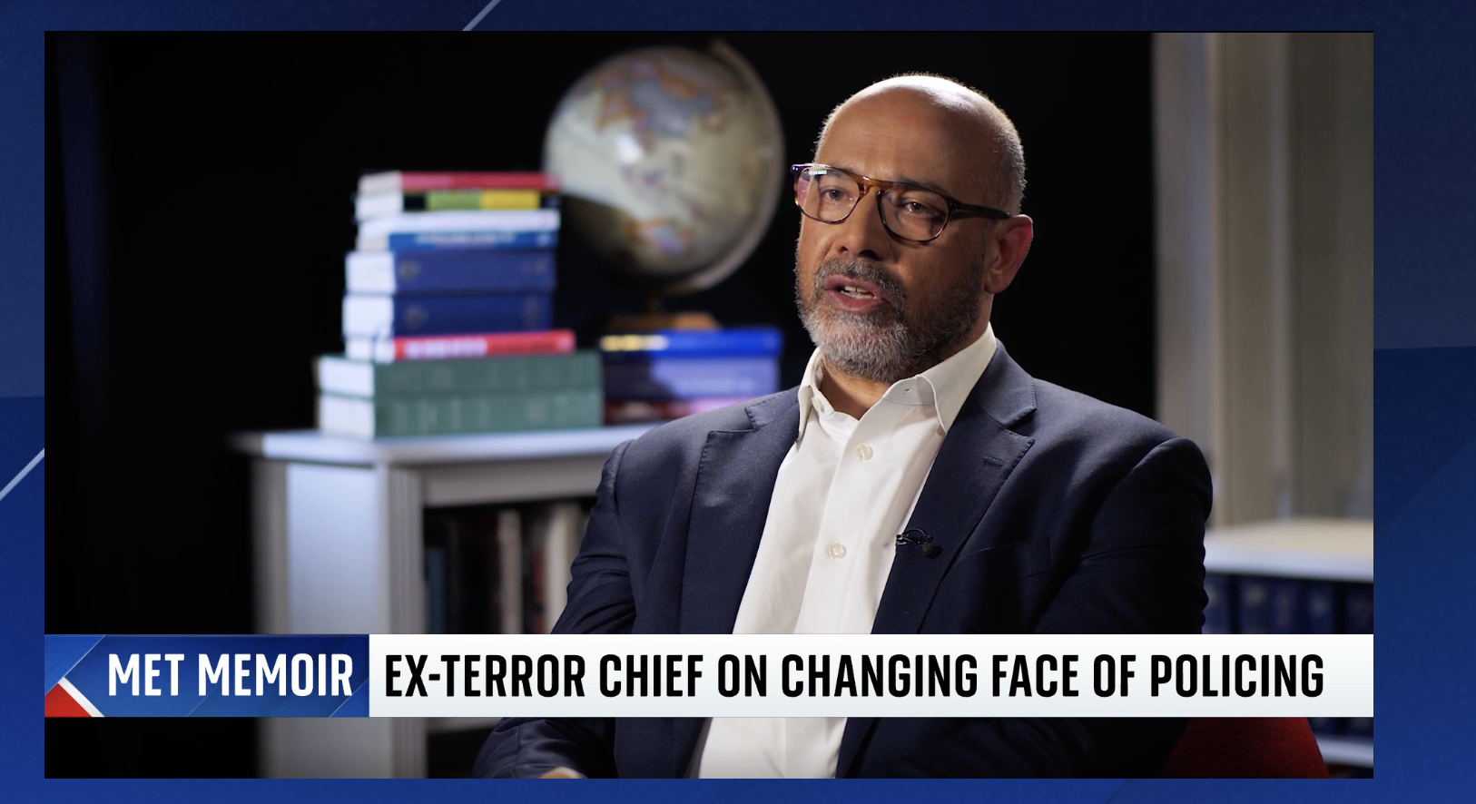 A man in a suit and glasses being interviewed, with a title banner reading 'MET Memoir: Ex-Terror Chief on Changing Face of Policing' in an office with a globe and stacked books in the background.