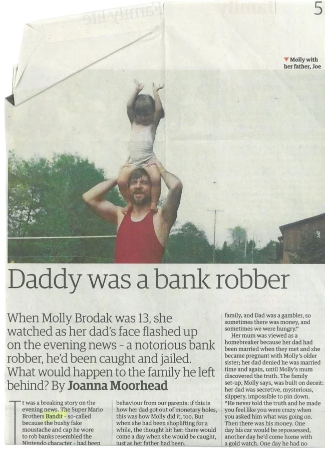 A man with a beard in a red tank top, carrying a young girl on his shoulders, lifting her hands in the air outdoors with trees in the background.