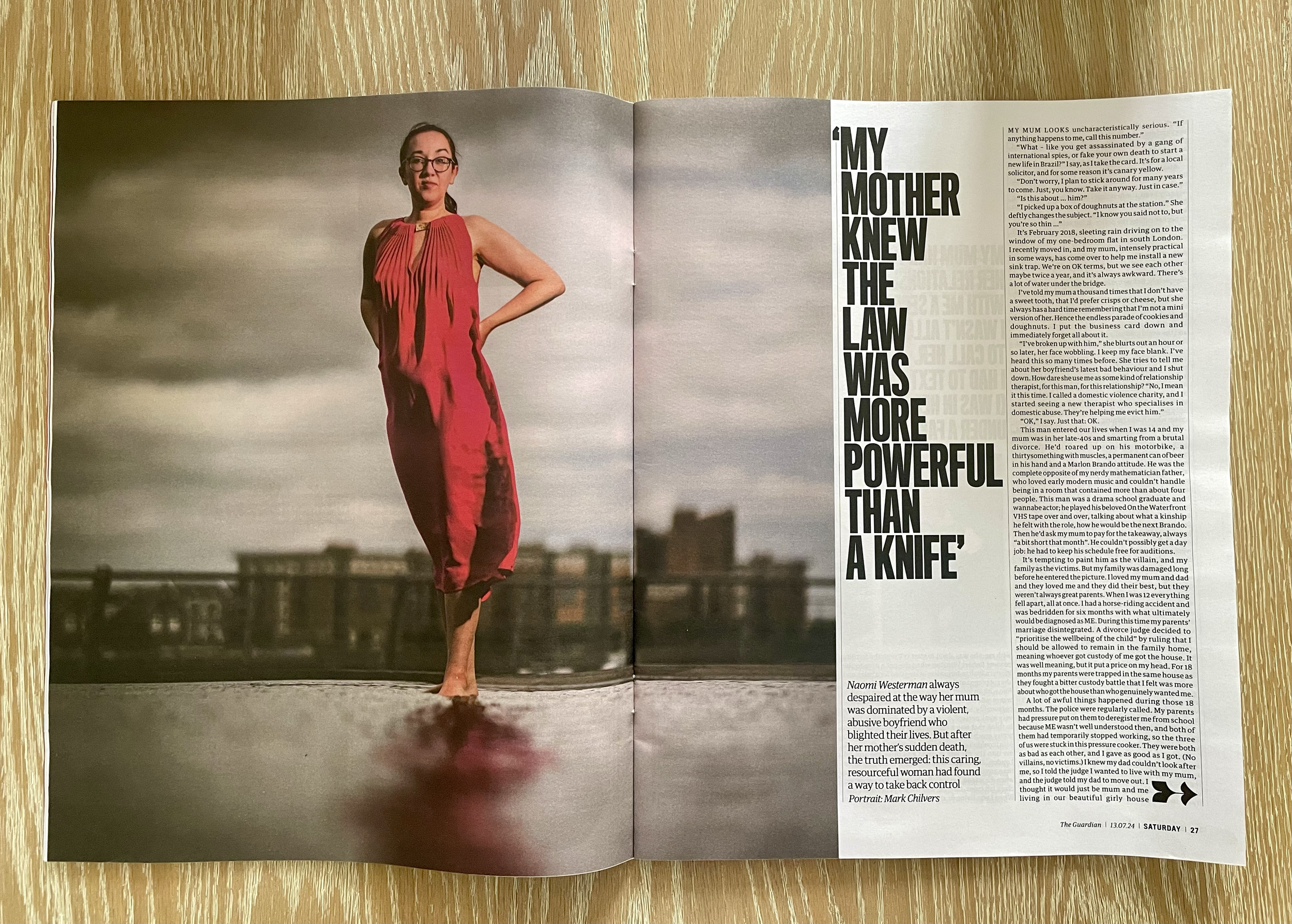 A woman standing outdoors on a rooftop with a city skyline in the background, wearing a red jumpsuit and glasses, with her left hand on her hip.