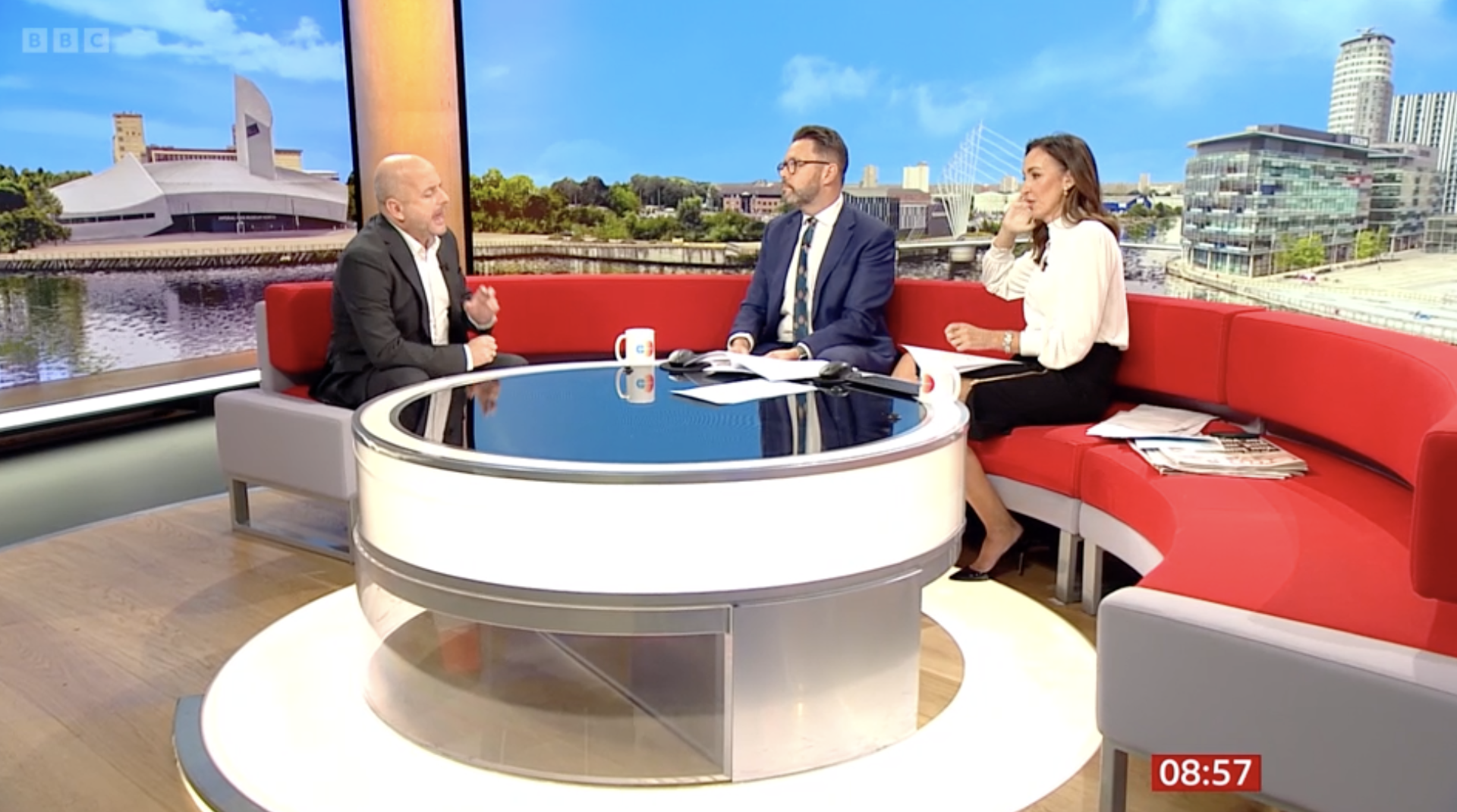 Three TV hosts sitting on a red curved sofa around a white and black round table in a studio with a cityscape backdrop. The man on the left, in a dark suit, is speaking; the man in the middle, in a blue suit with glasses, is listening; the woman on t