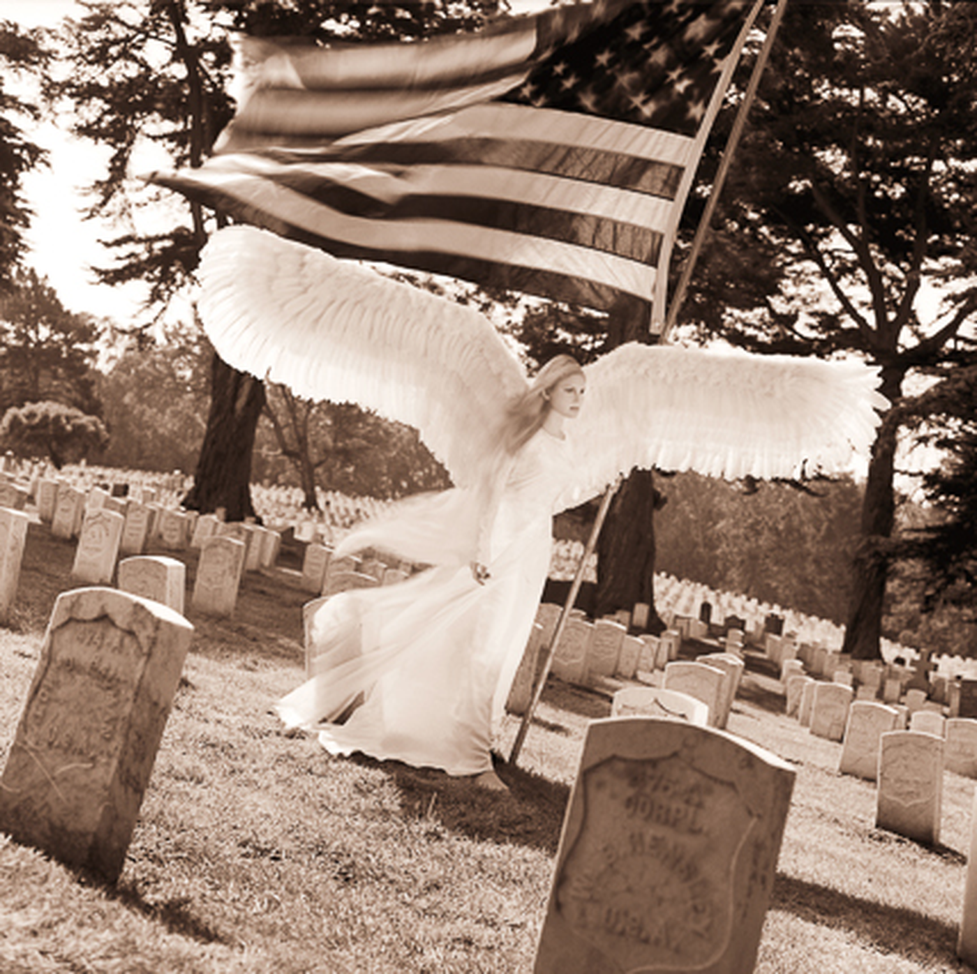 A woman dressed as an angel with large white wings standing in a cemetery, holding an American flag. Sepia tone.