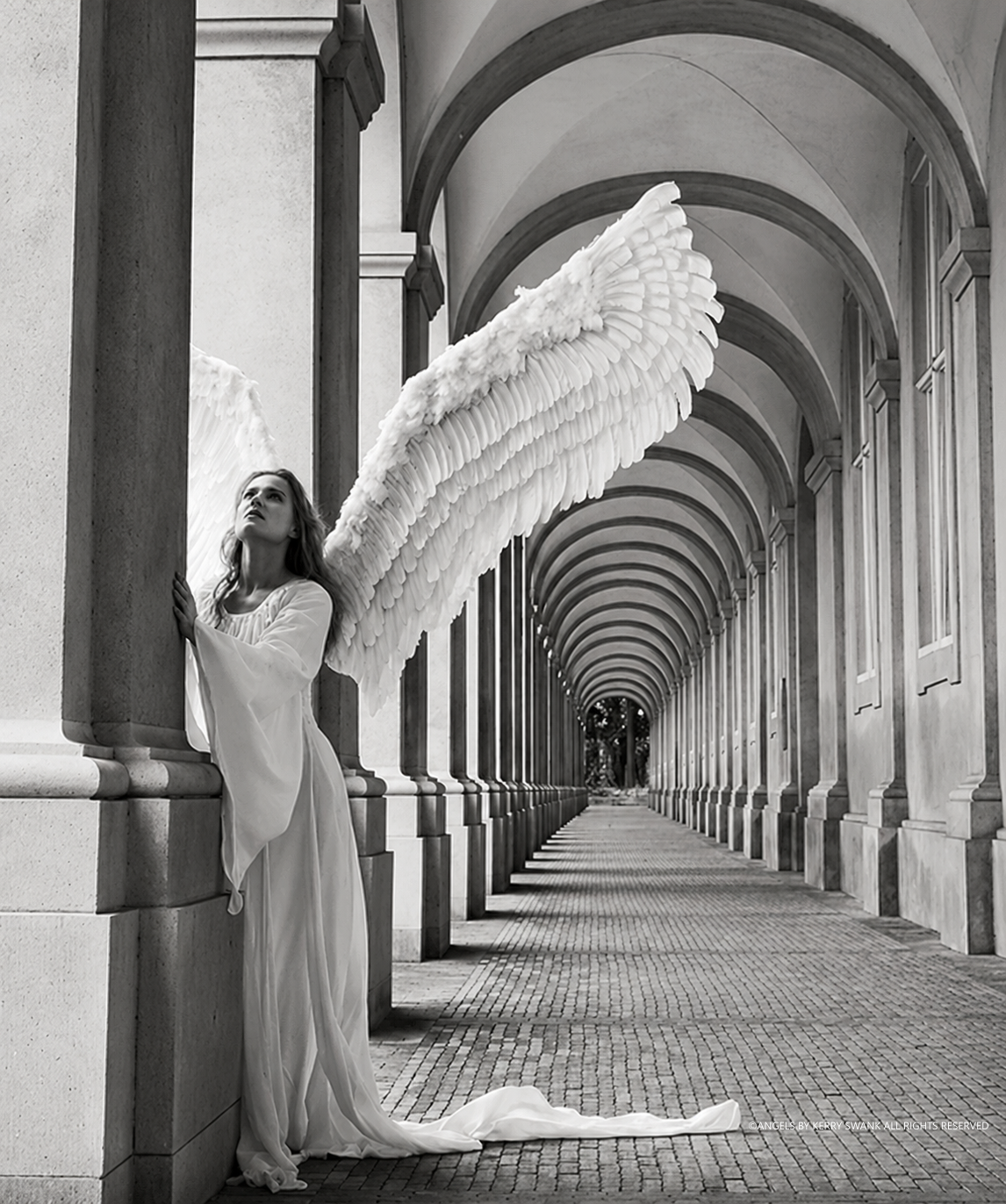 A woman with angel wings standing in a colonnade, looking upward with her hands resting on a column, in black and white.