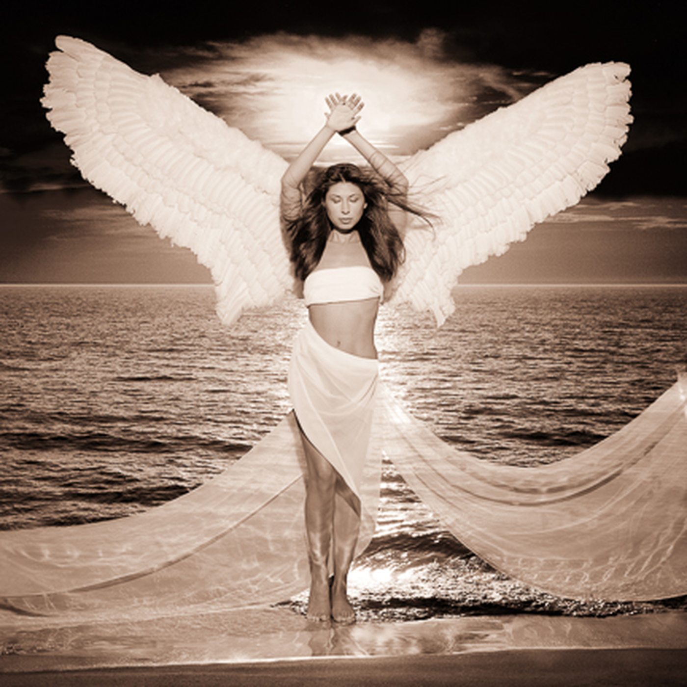 A woman in a white skirt and crop top with large angel wings standing on a dock by the ocean during sunset.