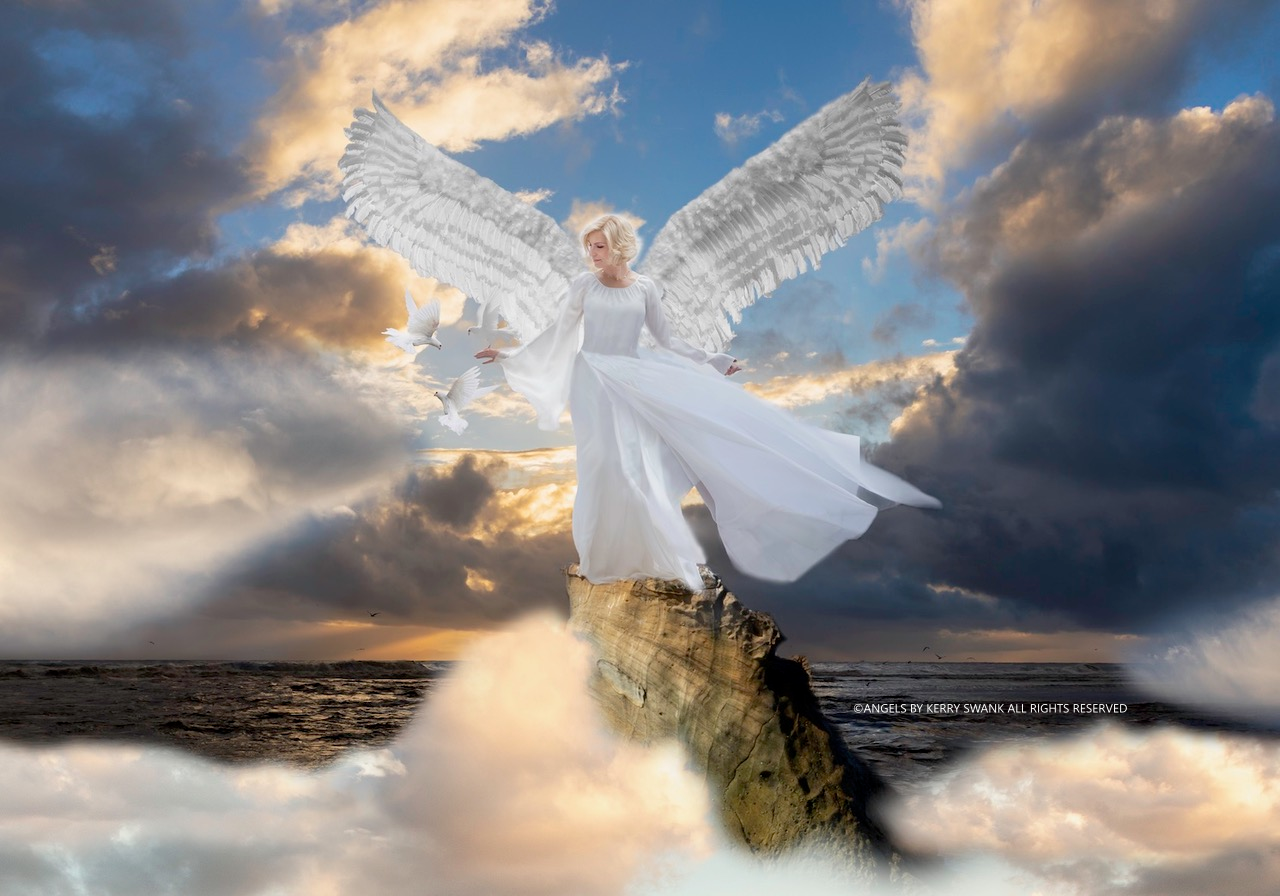 A woman dressed as an angel, with large white wings, standing on a rock in the ocean during a sunset or sunrise, with clouds and flying doves around her.