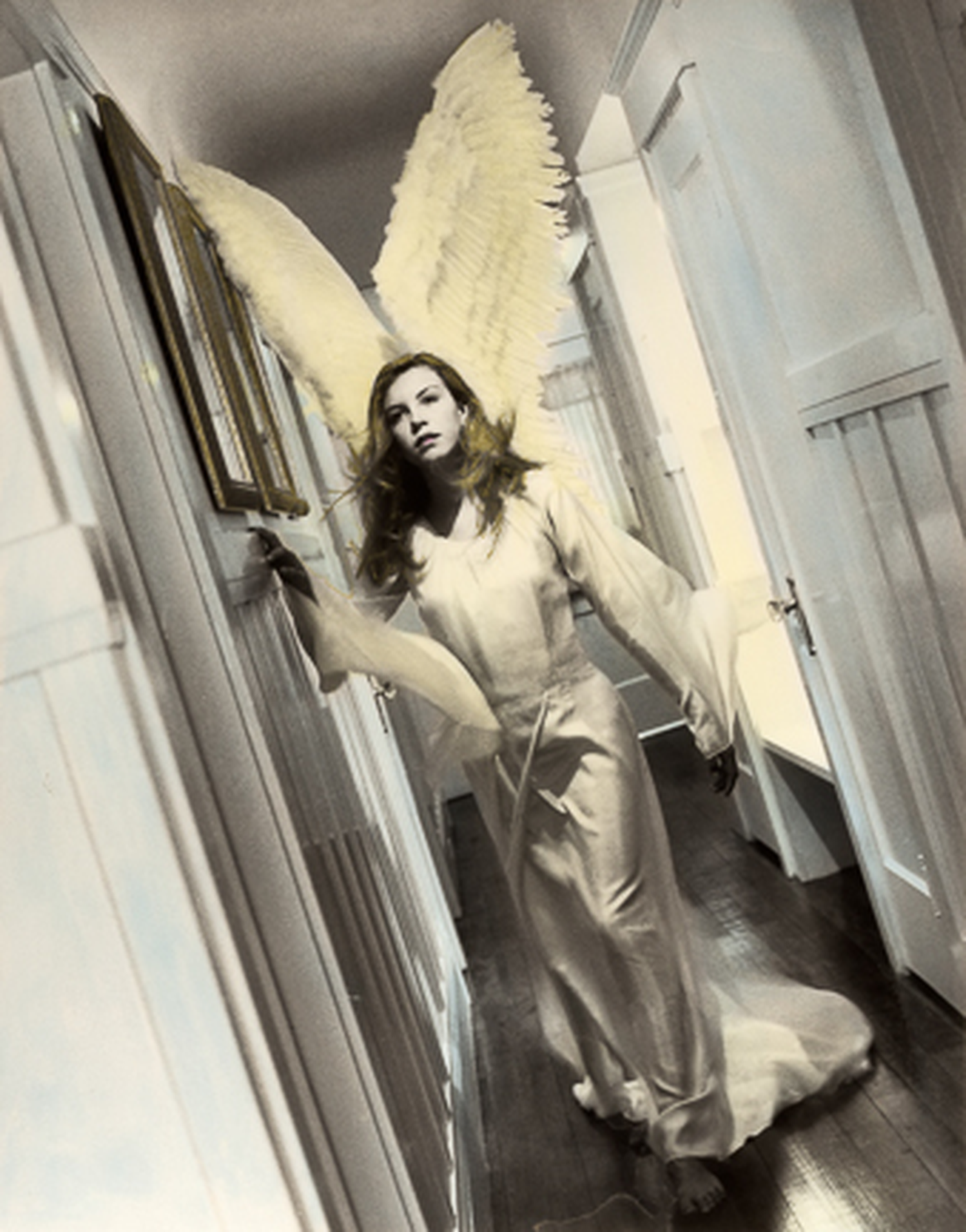 A person dressed as an angel with large yellow wings and a white gown, standing in a hallway with white door frames and panels, with long hair and an intense expression.