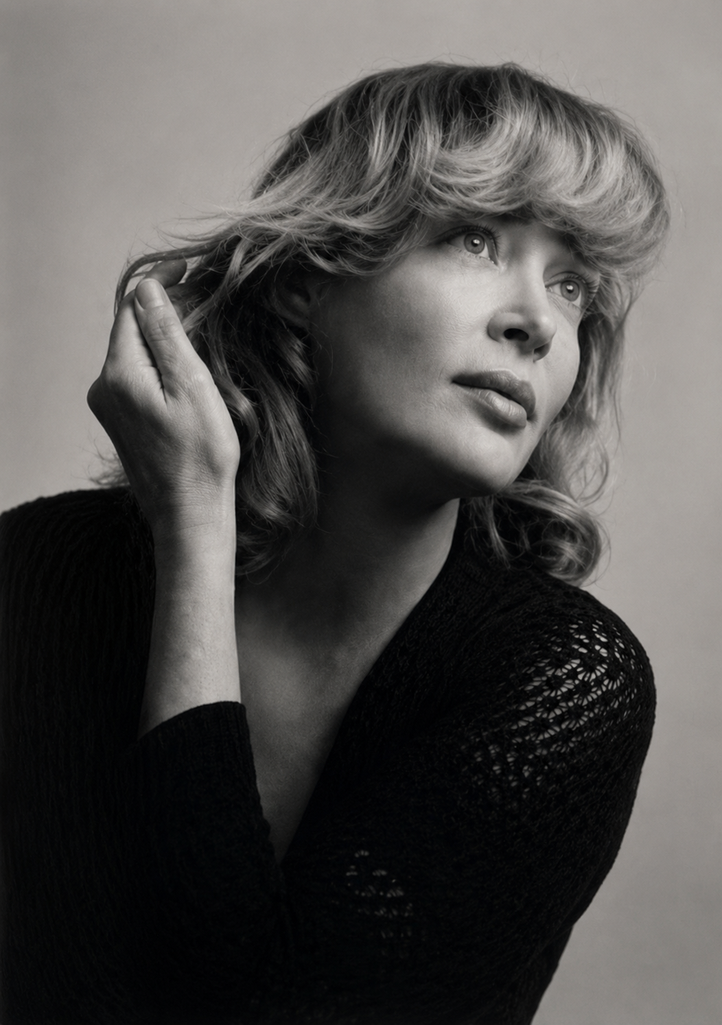Black and white portrait of a woman with wavy, shoulder-length hair, wearing a knitted black top, gazing thoughtfully to the side, with one hand near her hair.