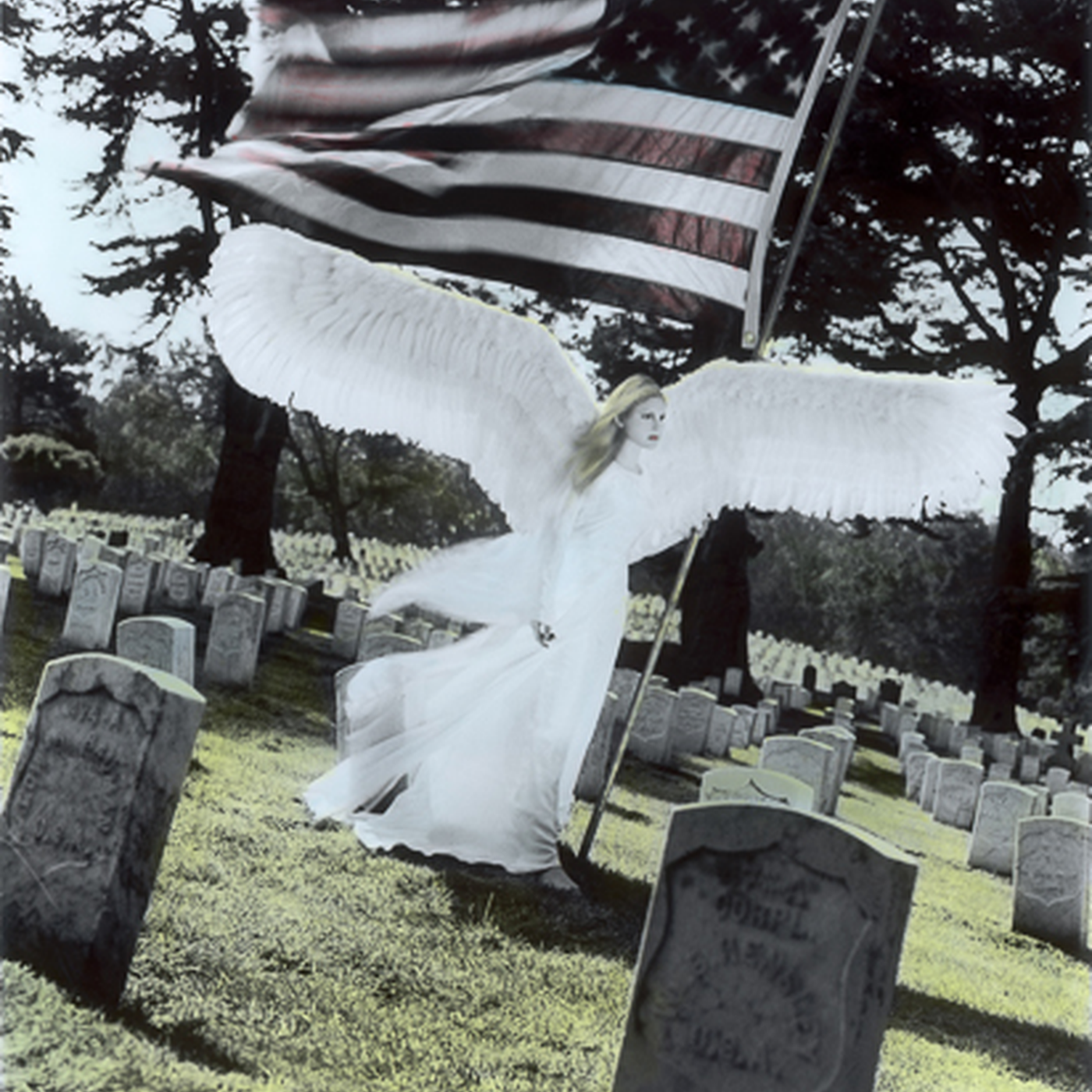 A female angel statue with white wings holding a large American flag, standing in a cemetery among many tombstones, with trees in the background.
