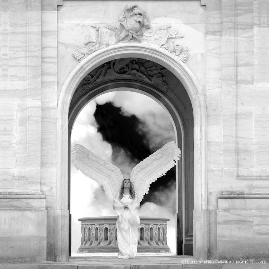 A woman dressed as an angel with large feathered wings and a flowing white gown standing beneath a grand archway with detailed carvings, looking up with her hands clasped, and a dark cloudy sky in the background.