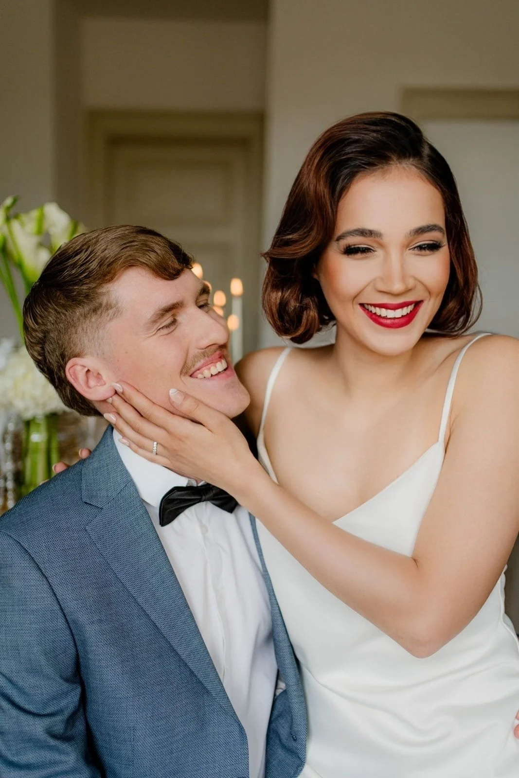 Ein lachendes Paar, bei einer Hochzeit, mit der Frau, die den Mann liebevoll ansieht, in eleganter Kleidung, im Hintergrund Blumen und Kerzen.