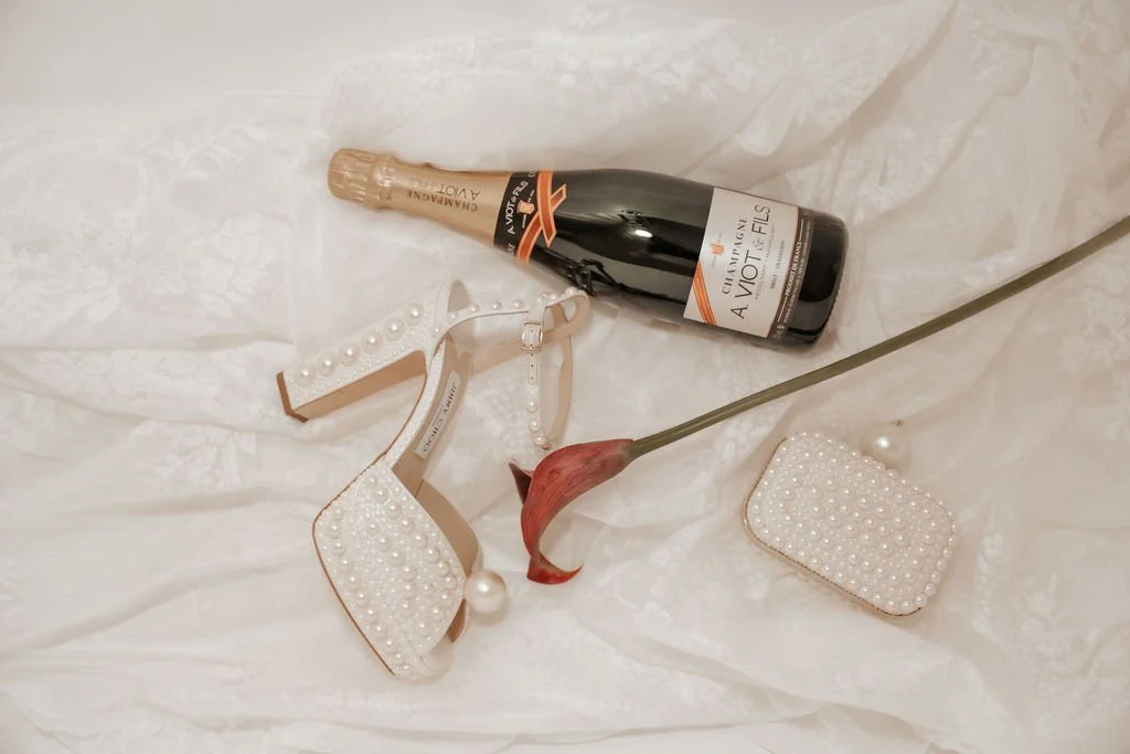 Sektflasche, weißes High Heels mit Perlen, eine rote Calla-Lilie, eine kleine Perlen-Handtasche, eine Perlenkette