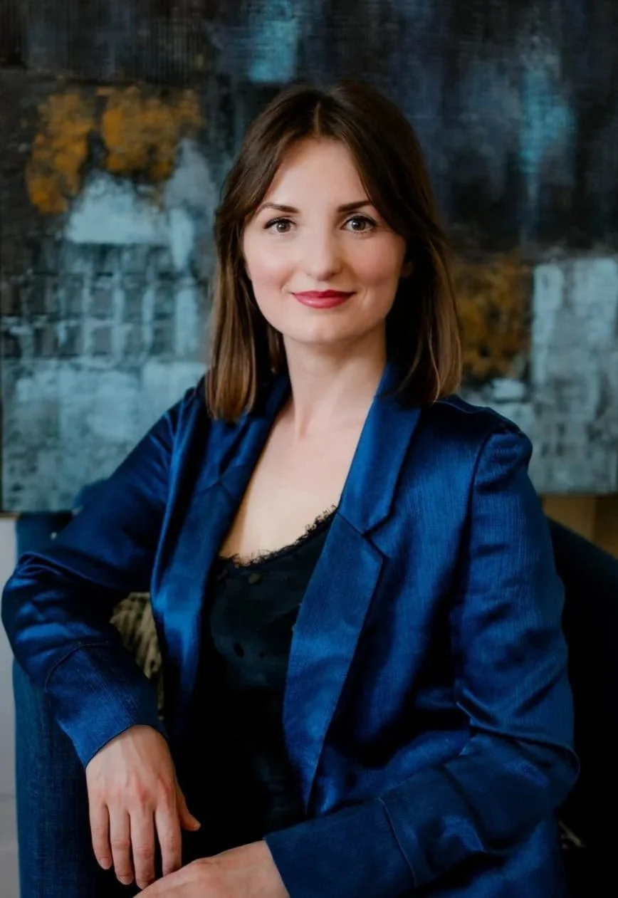 Portrait einer lachenden Frau mit braunen Haaren, in blauer Jacke, im Hintergrund ein abstraktes Gemälde.