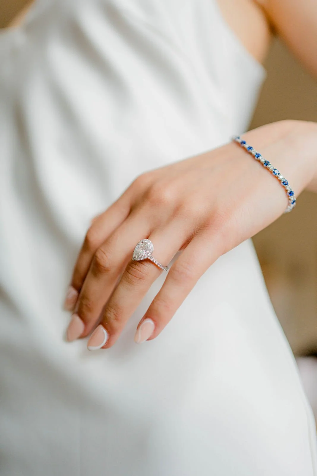 Nahaufnahme einer Hand mit einem Verlobungsring und einem Armband, vor einem weißen Kleid.