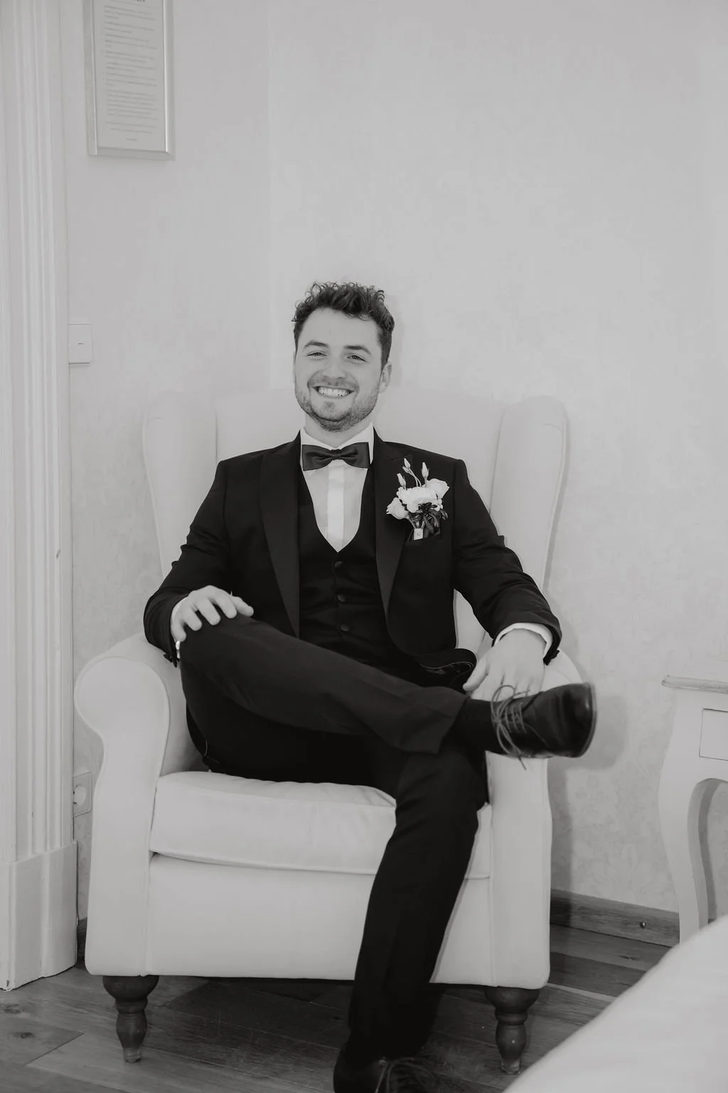 Junger Mann in formeller Kleidung, sitzend auf einem Sessel, lachend. Er trägt einen schwarzen Anzug, eine Fliege und ein Blumenboutonnière am Revers, vermutlich an einem besonderen Anlass wie einer Hochzeit.