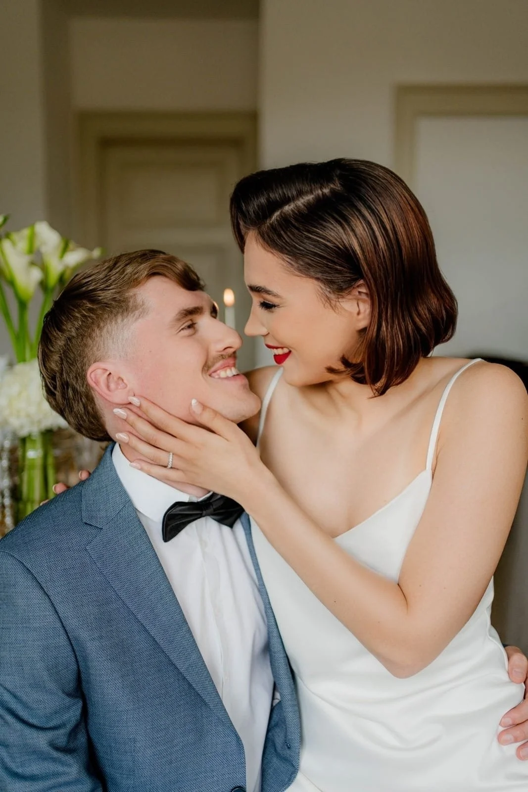 Ein glückliches Paar bei einer Hochzeit, der Mann trägt einen blauen Anzug mit Fliege, die Frau trägt ein weißes Brautkleid, sie schauen sich verliebt an