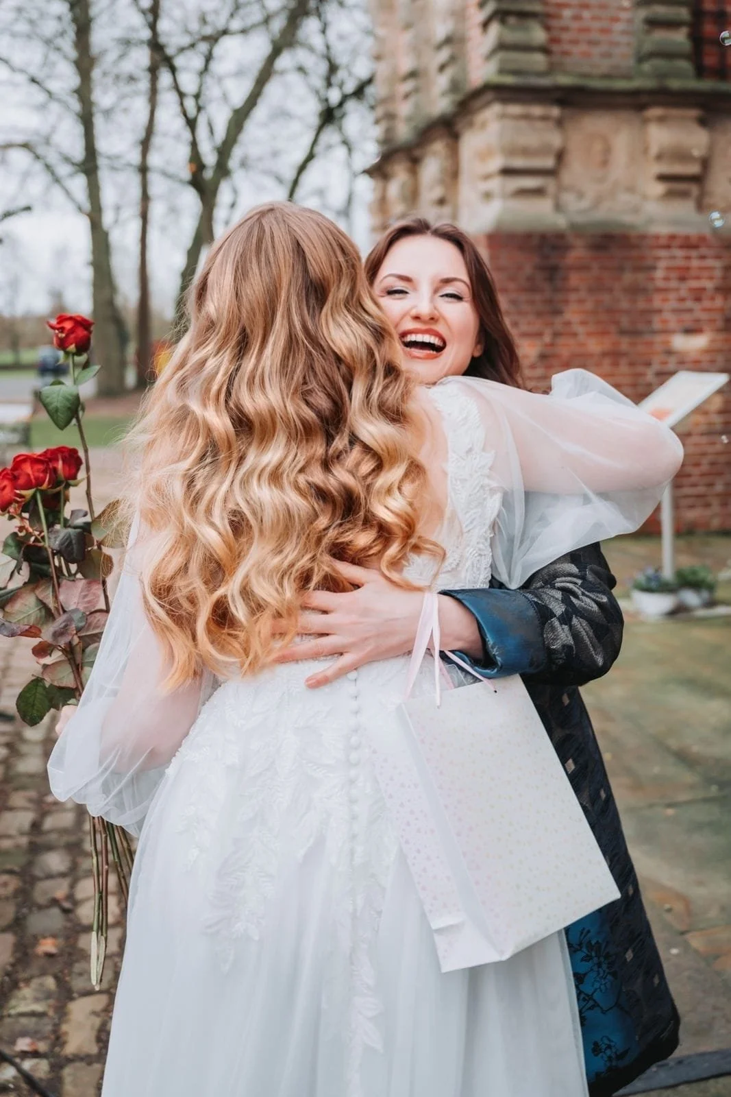 Zwei Frauen umarmen sich, eine in einem weißen Hochzeitskleid mit lockigem, roten Haar, die andere in dunkler Kleidung, beide lachen bei einem draußen Treffen.