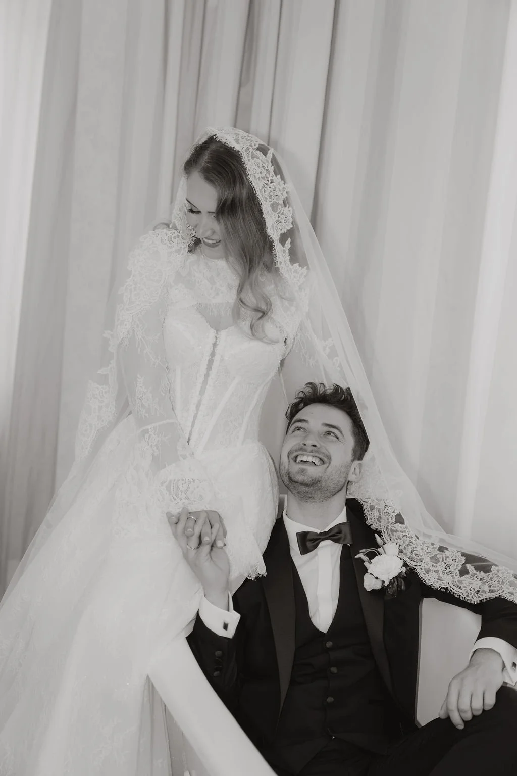 Braut und Bräutigam im Hochzeitskleid, lachend und Händchen haltend, in einem Raum mit Holzvertäfelung, schwarzer Anzug, geflochtener Haarschopf, Blumenkorsage, Hochzeitskleid mit Spitze, weiße Schleier.