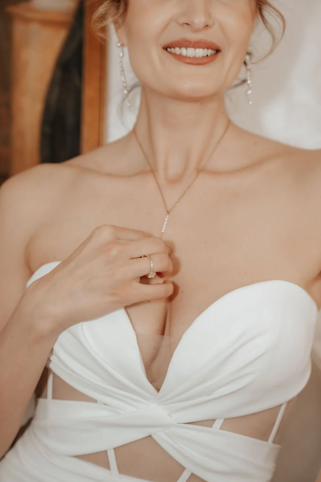 Nahaufnahme eines lachenden Mädchens in weißem Kleid, mit Schmuck und einer Hand an der Brust, im Hintergrund Holzmöbel.