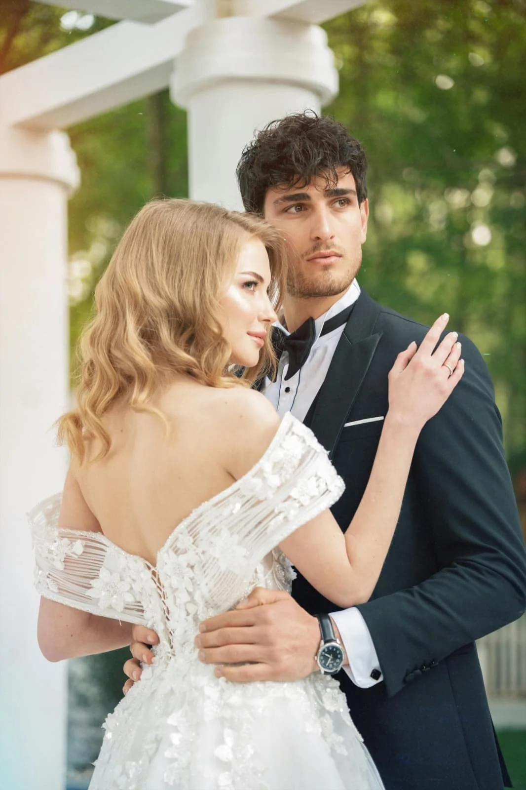 Ein Brautpaar in formeller Kleidung steht im Freien, umgeben von grünen Bäumen, während sie sich aneinander lehnen.